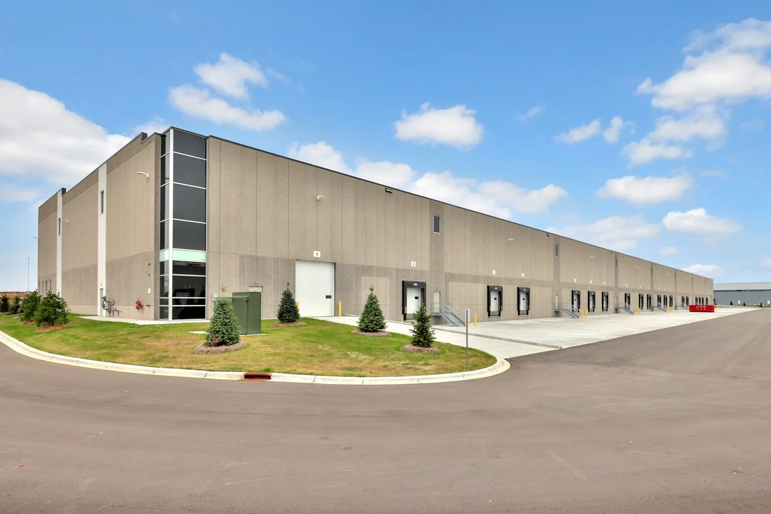 Large warehouse building with multiple loading docks, surrounded by a paved lot and small landscaped trees under a partly cloudy sky.