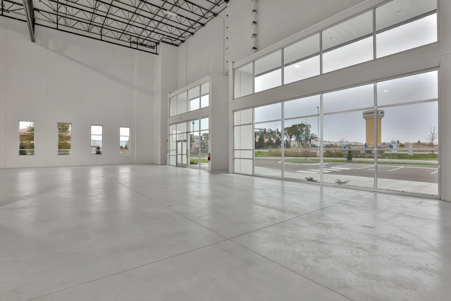 Empty commercial space with large glass windows and high ceiling, view of parking lot and water tower outside.