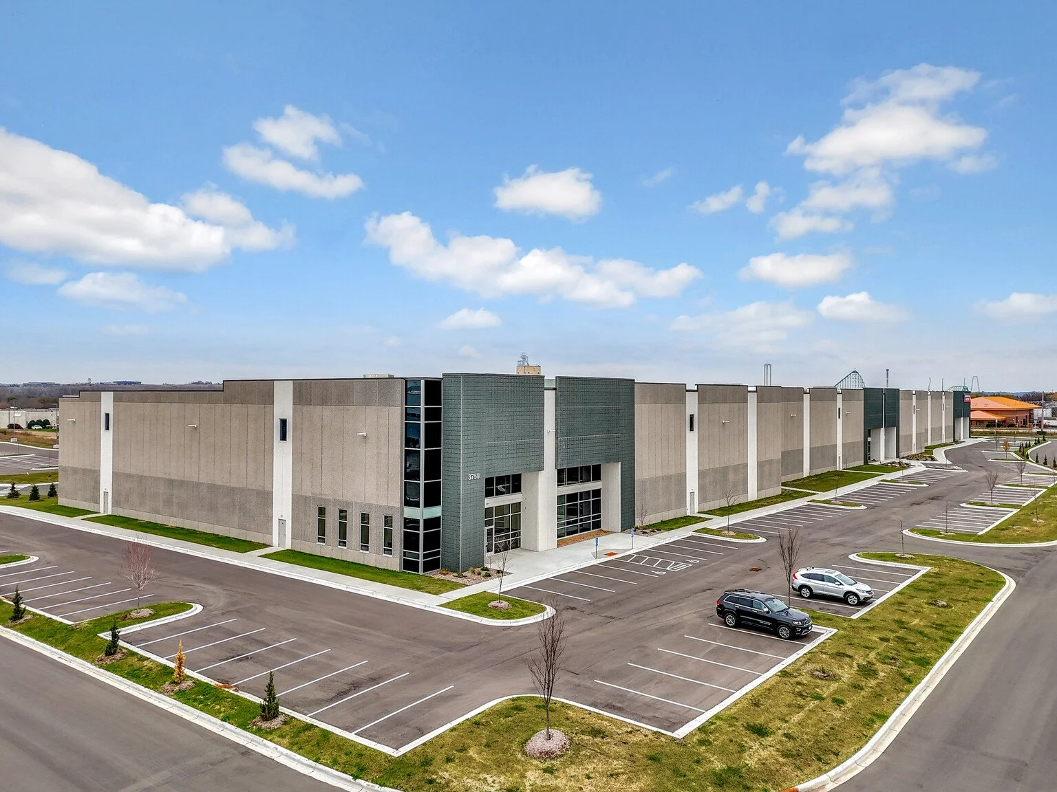 Large industrial or commercial building with parking lot and a few cars, surrounded by green grass and leafless trees under a blue sky with scattered clouds.