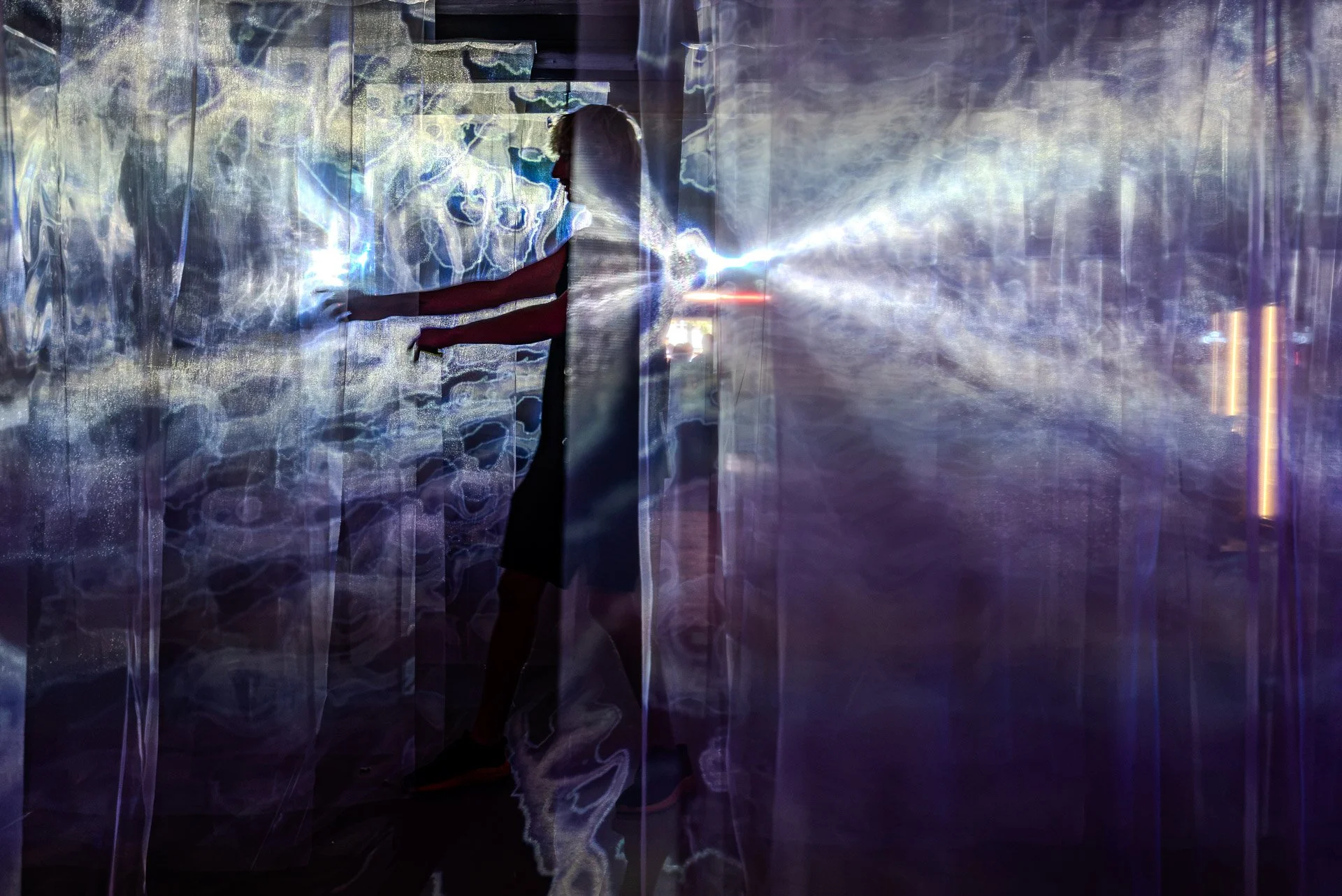 A person is inside an illuminated, reflective, or holographic space with abstract, colorful light patterns, creating a surreal and futuristic atmosphere.