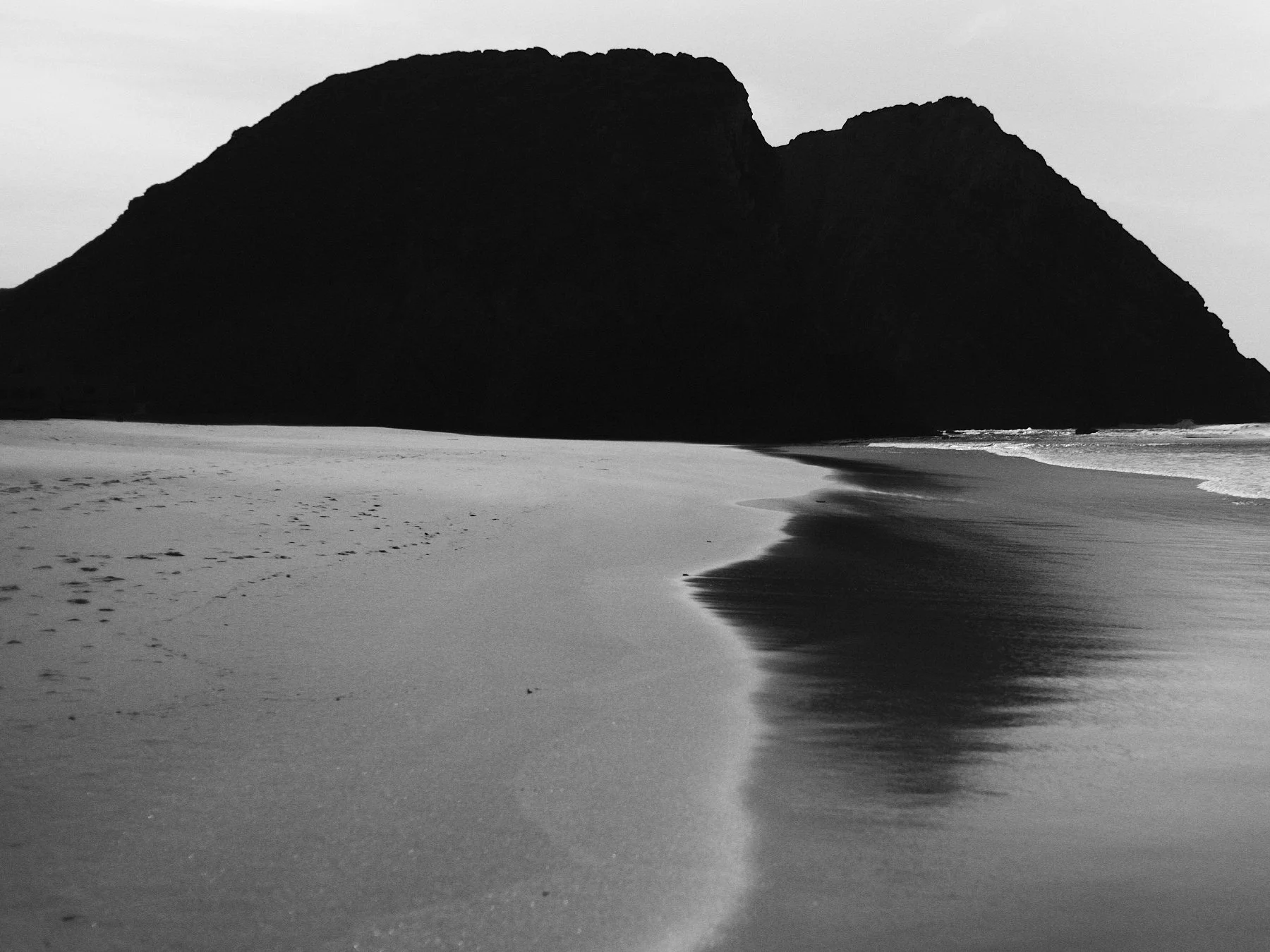 Black and white photo of a beach with a large mountain or cliff in the background, shoreline with gentle waves, and footprints in the sand.