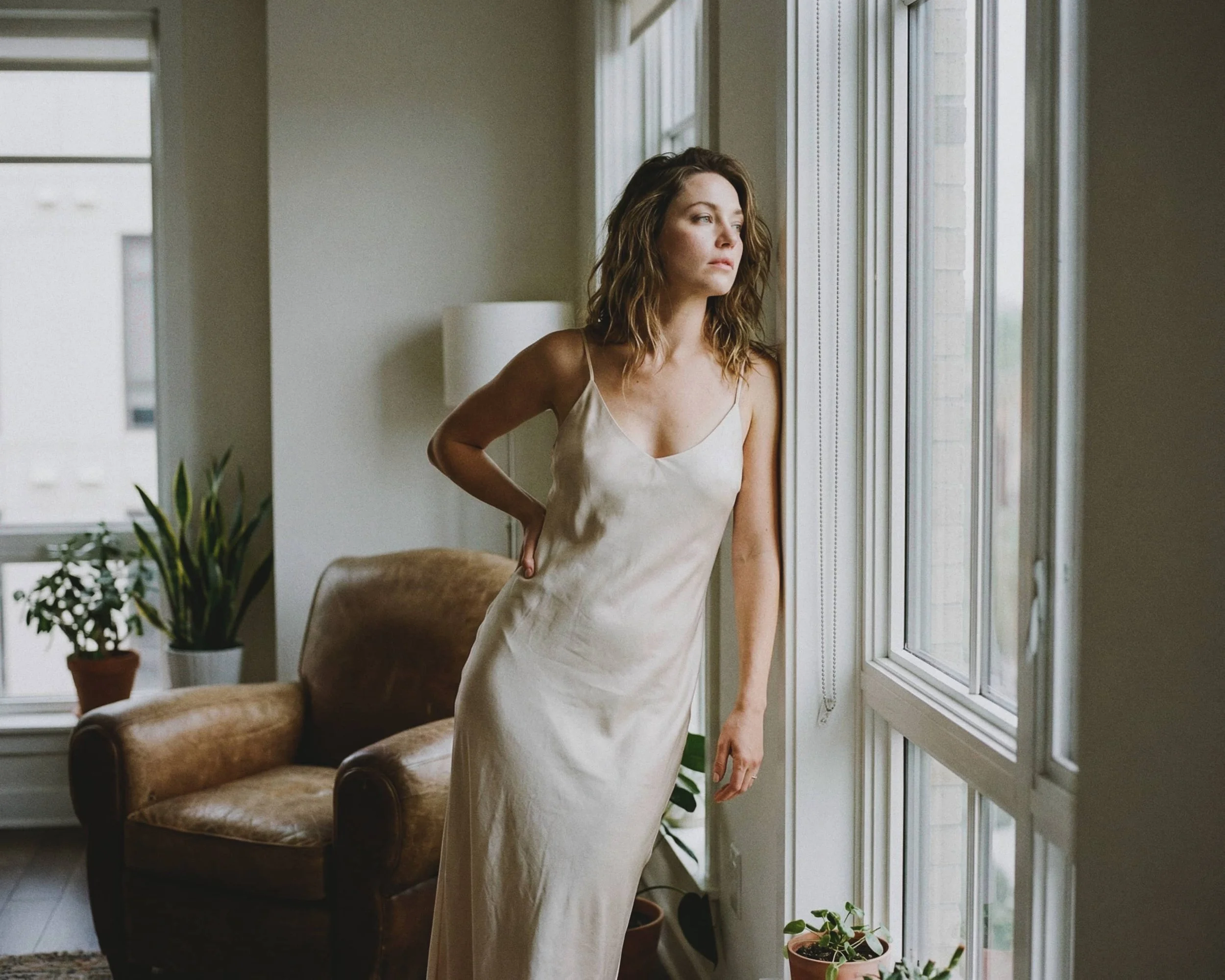 Mujer con vestido largo de seda, mirando por la ventana, en una sala con plantas y un sillón de cuero