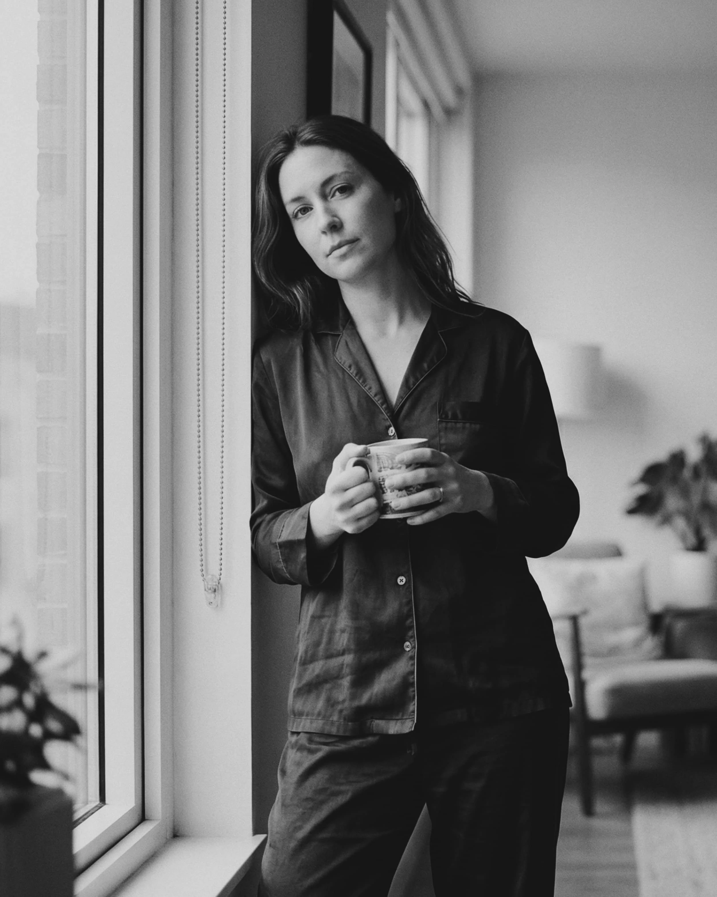 Mujer con pijama de satén negro sosteniendo una taza de cerámica, de pie junto a una ventana en un ambiente interior moderno.