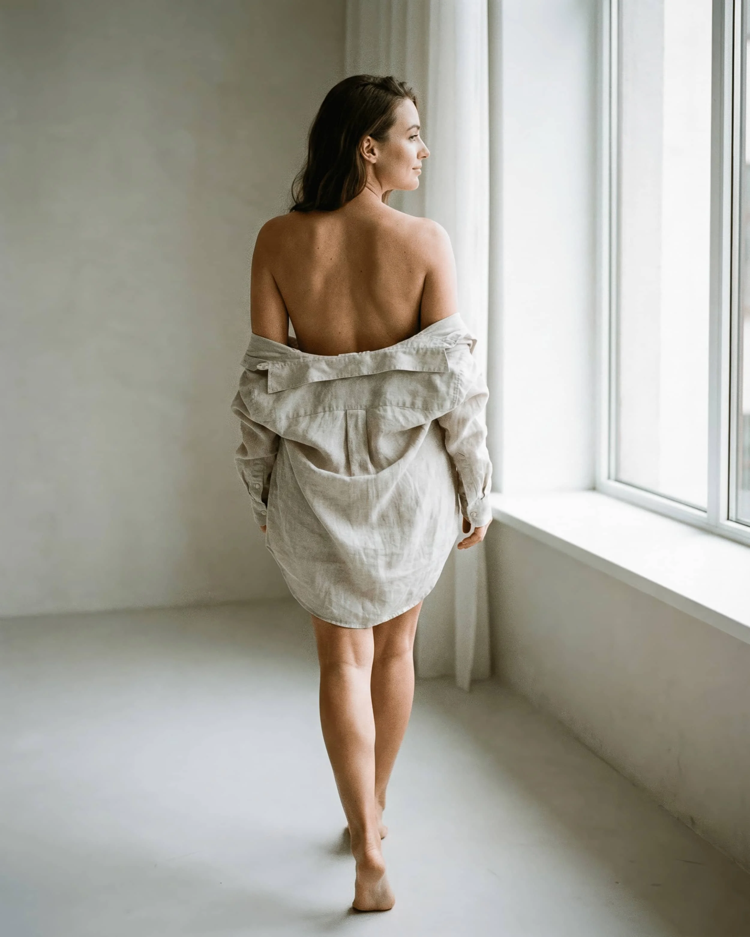 Mujer desnuda mirando por la ventana en una habitación con paredes blancas y cortinas blancas