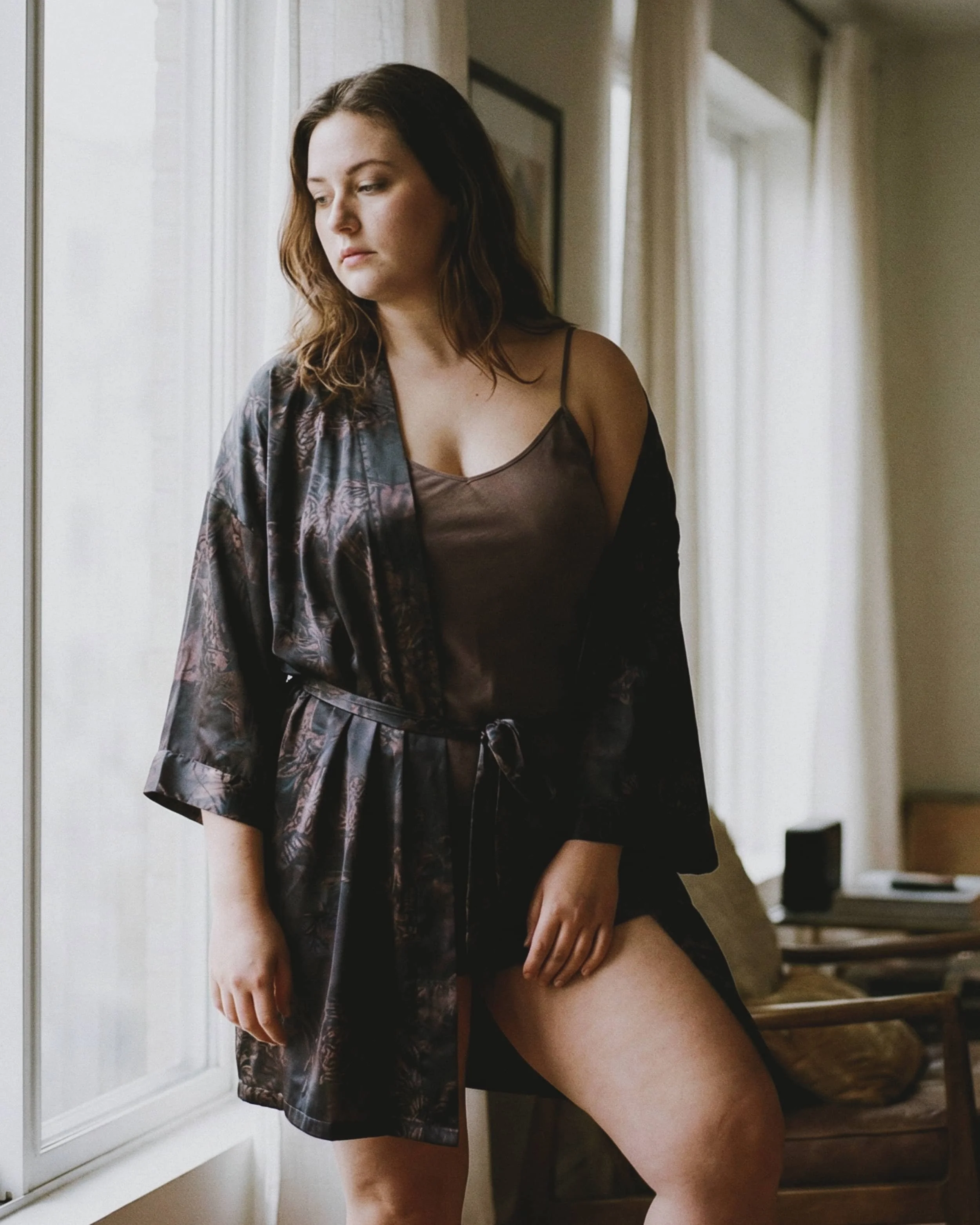 Mujer con vestido de camisón de seda y bata oscuro, de pie junto a una ventana en una habitación bien iluminada.