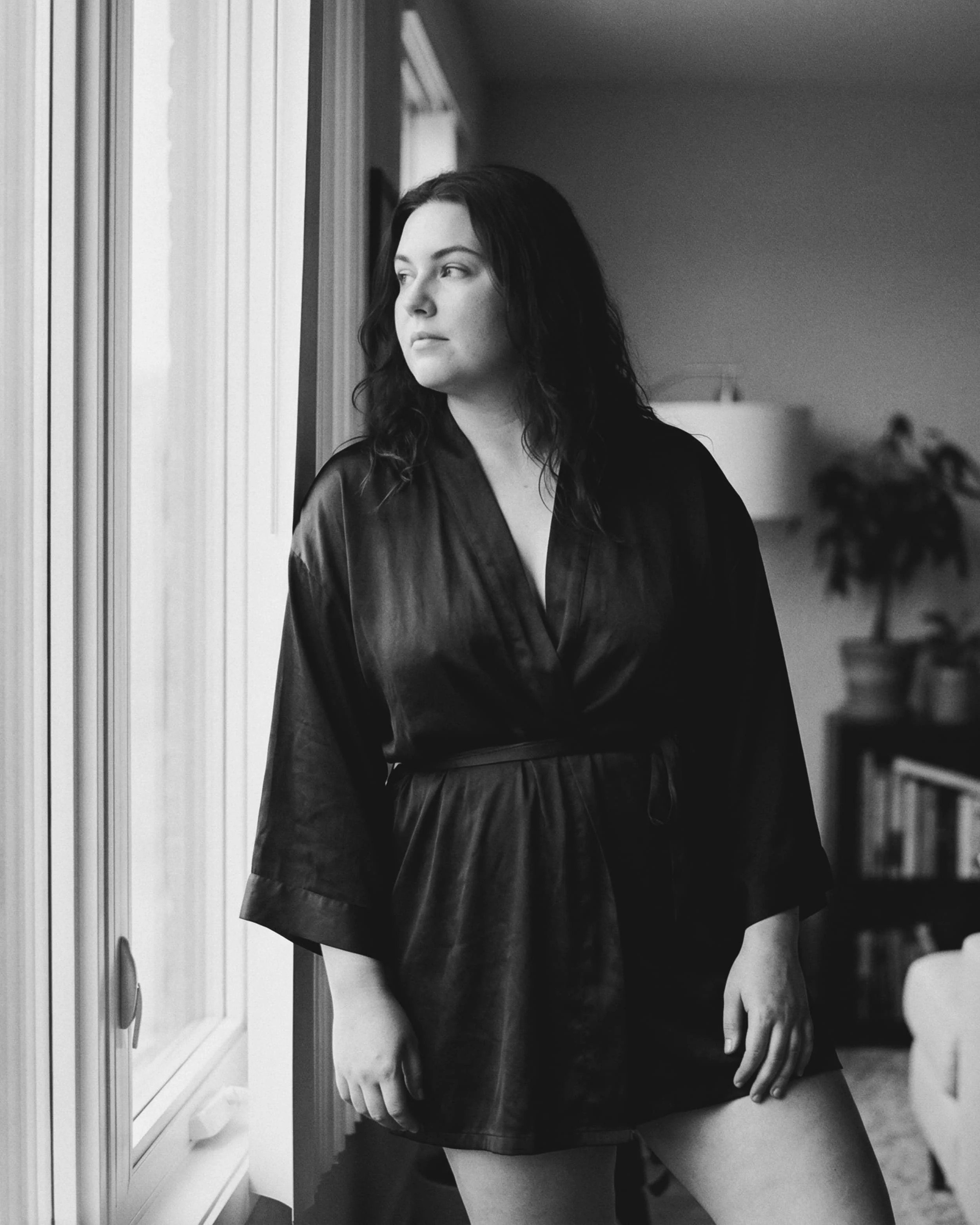Retrato en blanco y negro de una mujer de cabello oscuro, usando una bata de seda, mirando por la ventana en un interior con muebles y plantas.