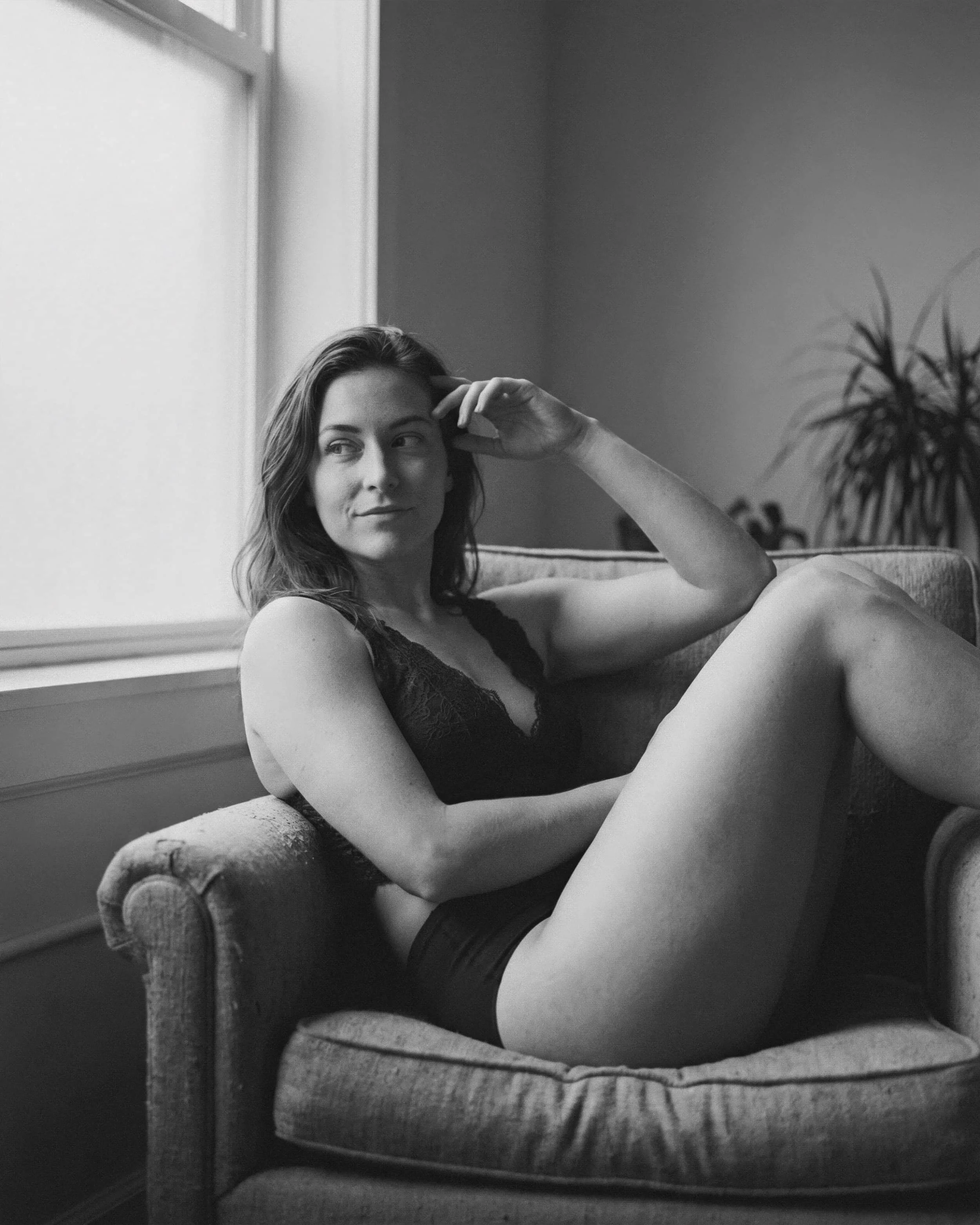 Mujer joven en ropa negra sentada en un sillón, con expresión pensativa, en una habitación luminosa.