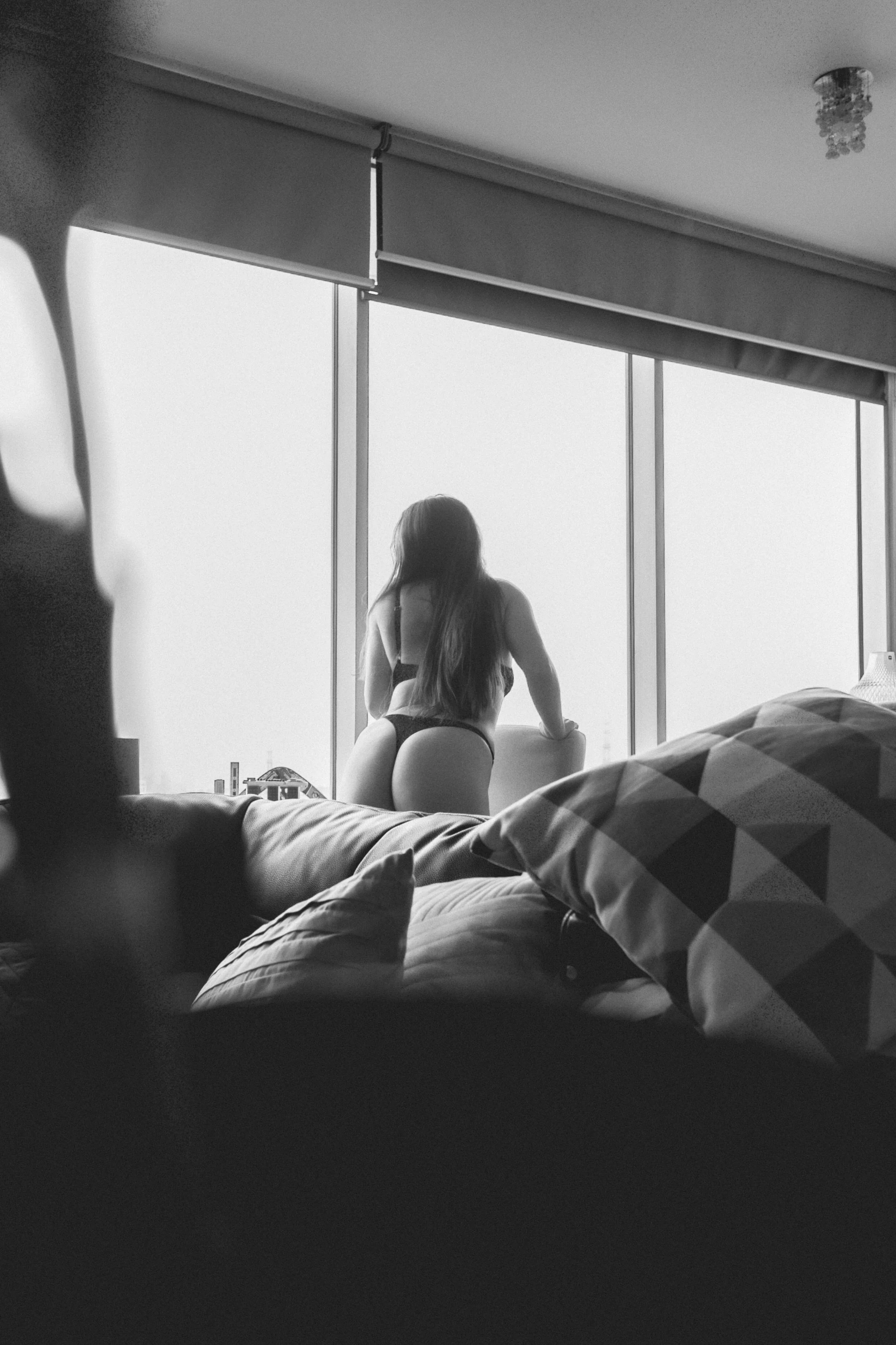 Mujer de espaldas con lencería negra mirando por una ventana grande en un apartamento, en blanco y negro.