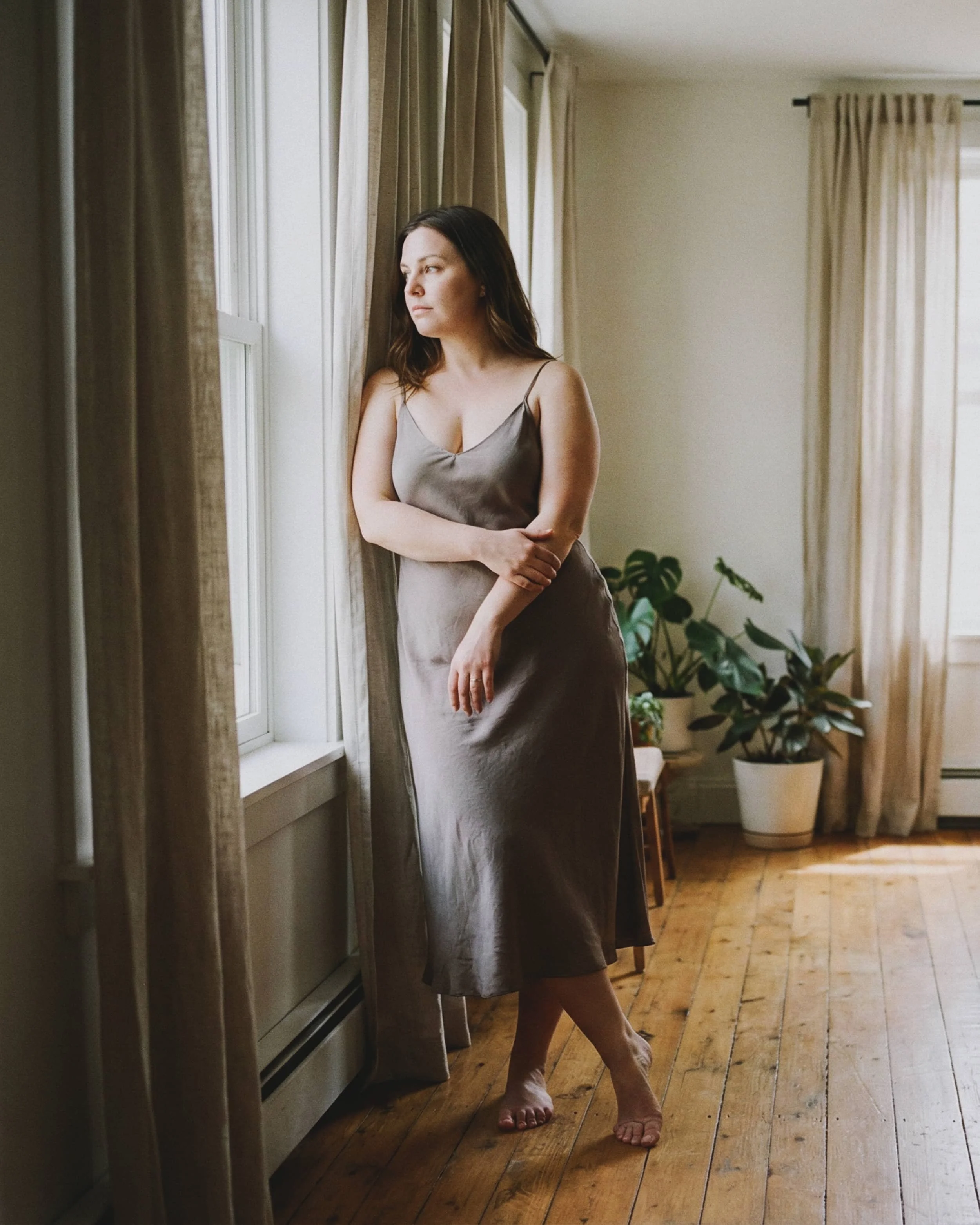 Mujer de cabello castaño con vestido satinado gris claro, de pie en una habitación con cortinas beige, plantas en macetas y ventanas con vista exterior, mirando pensativa por la ventana.