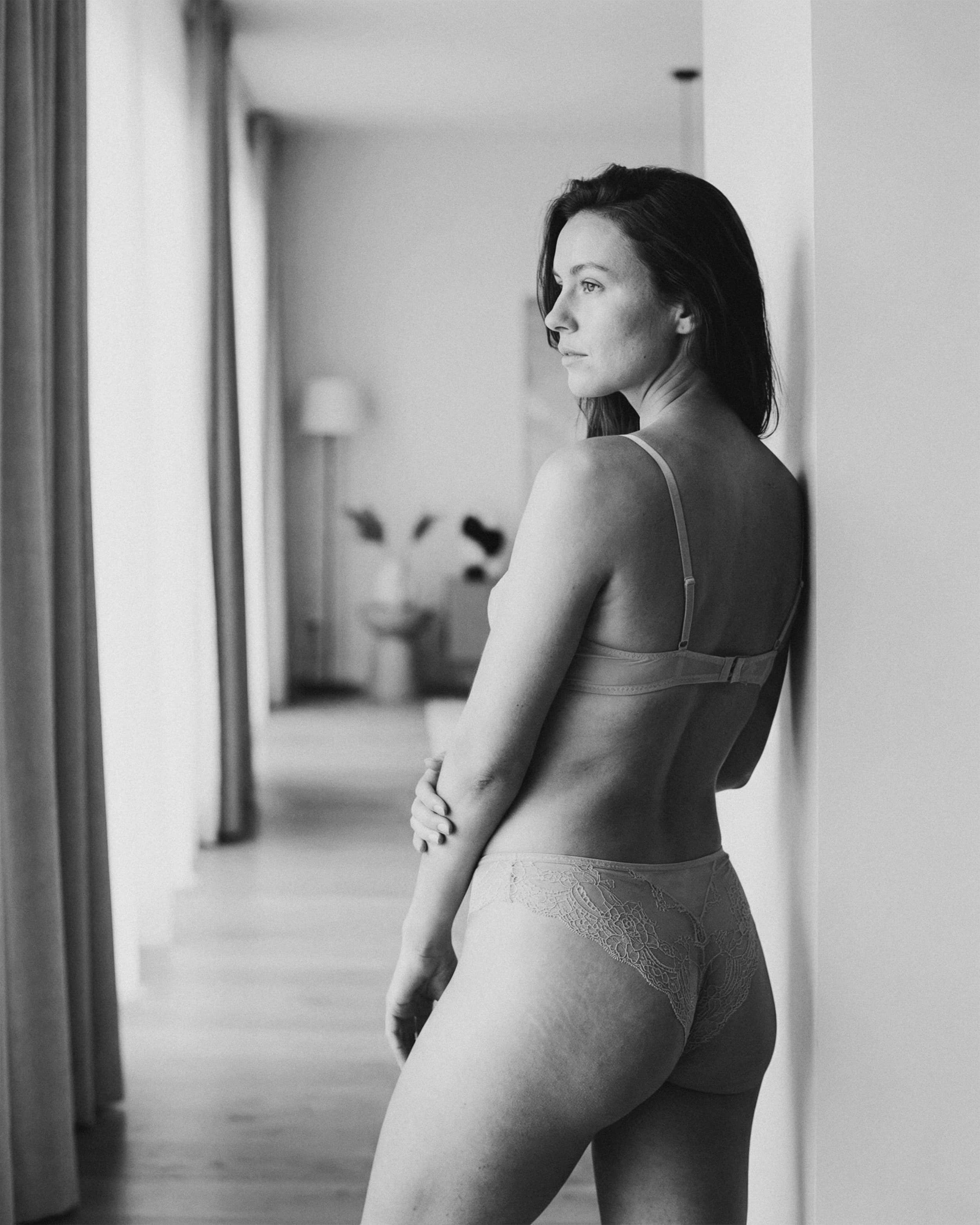 Mujer joven con lencería de encaje, de espaldas, en un pasillo, mirando hacia un lado, en blanco y negro.