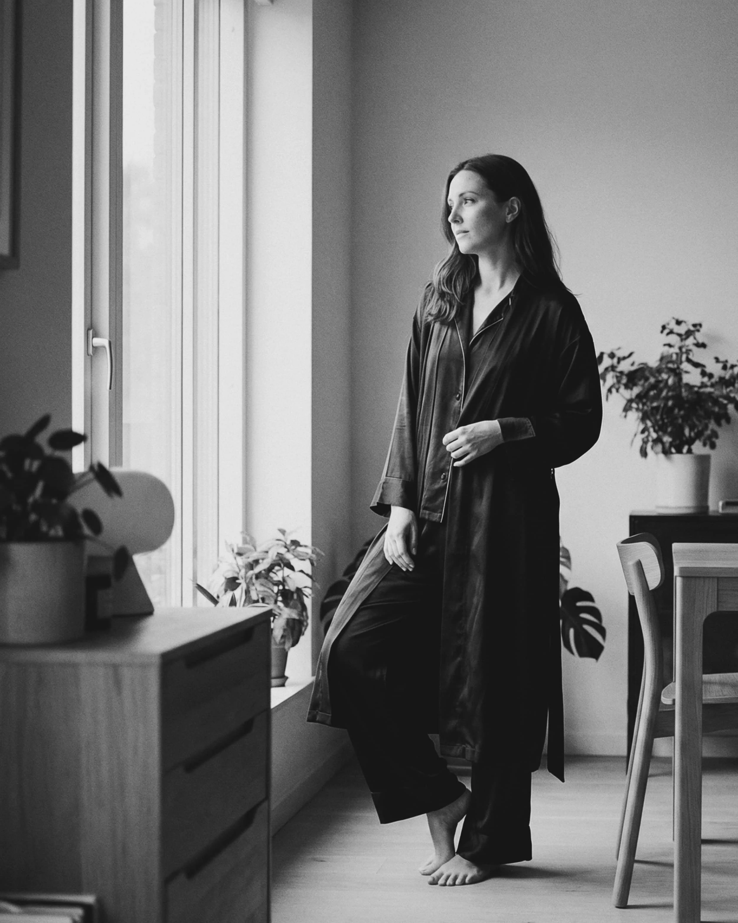 Mujer de cabello largo, de pie en una habitación con plantas, vestida con una túnica larga de seda, mirando por la ventana