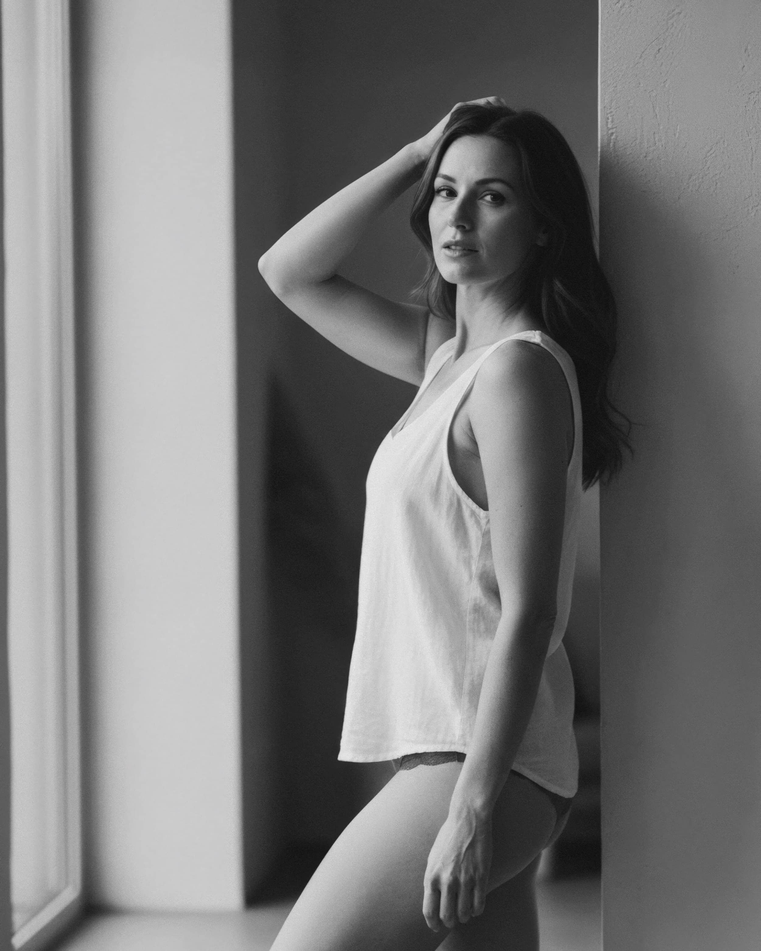 Mujer joven con cabello largo y oscuro, posa junto a la pared, lleva camiseta sin mangas y pantalones cortos, en una fotografía en blanco y negro.