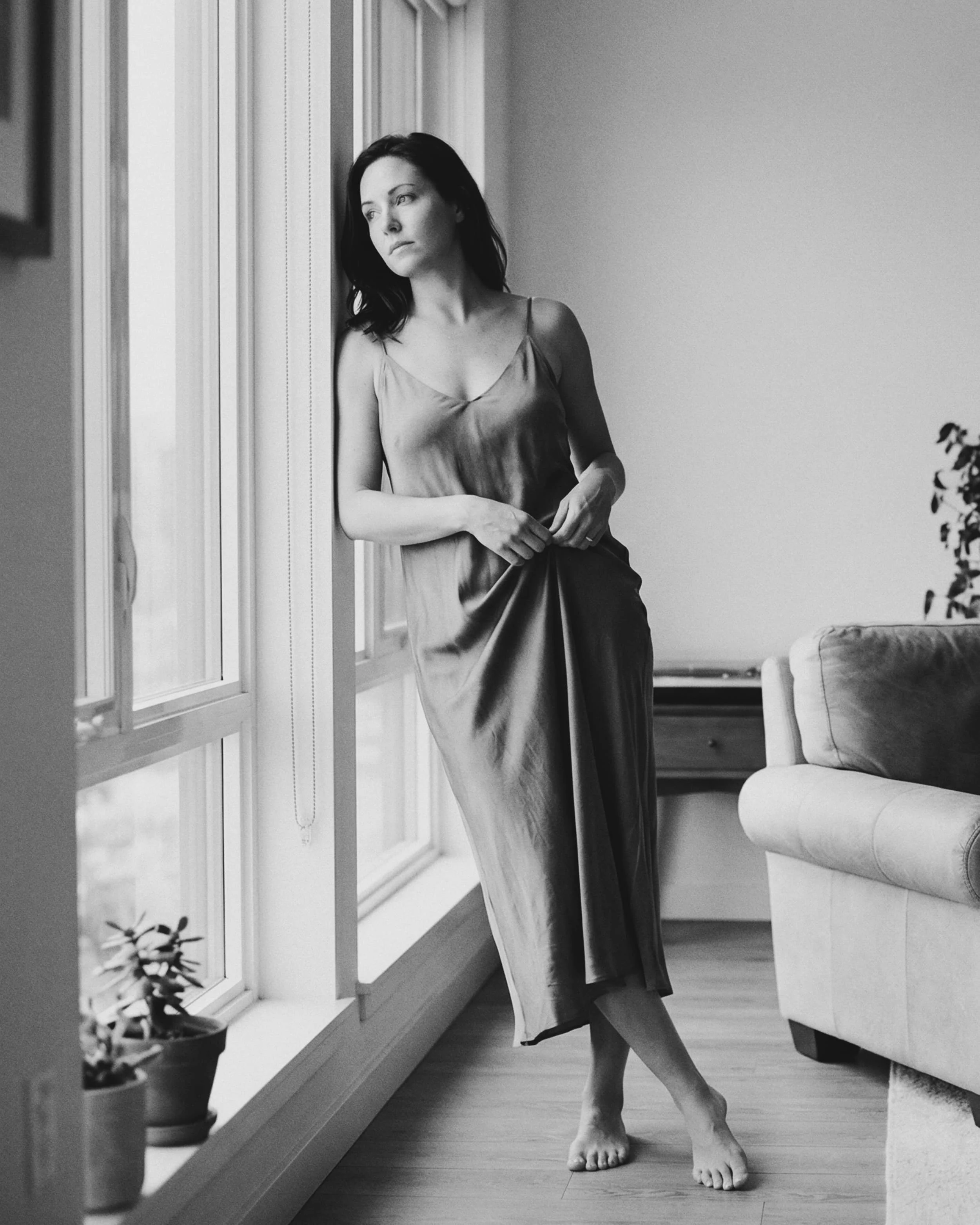 Mujer de cabello oscuro, vistiendo un vestido de seda sin mangas, de pie en una habitación junto a una ventana, con expresión pensativa.