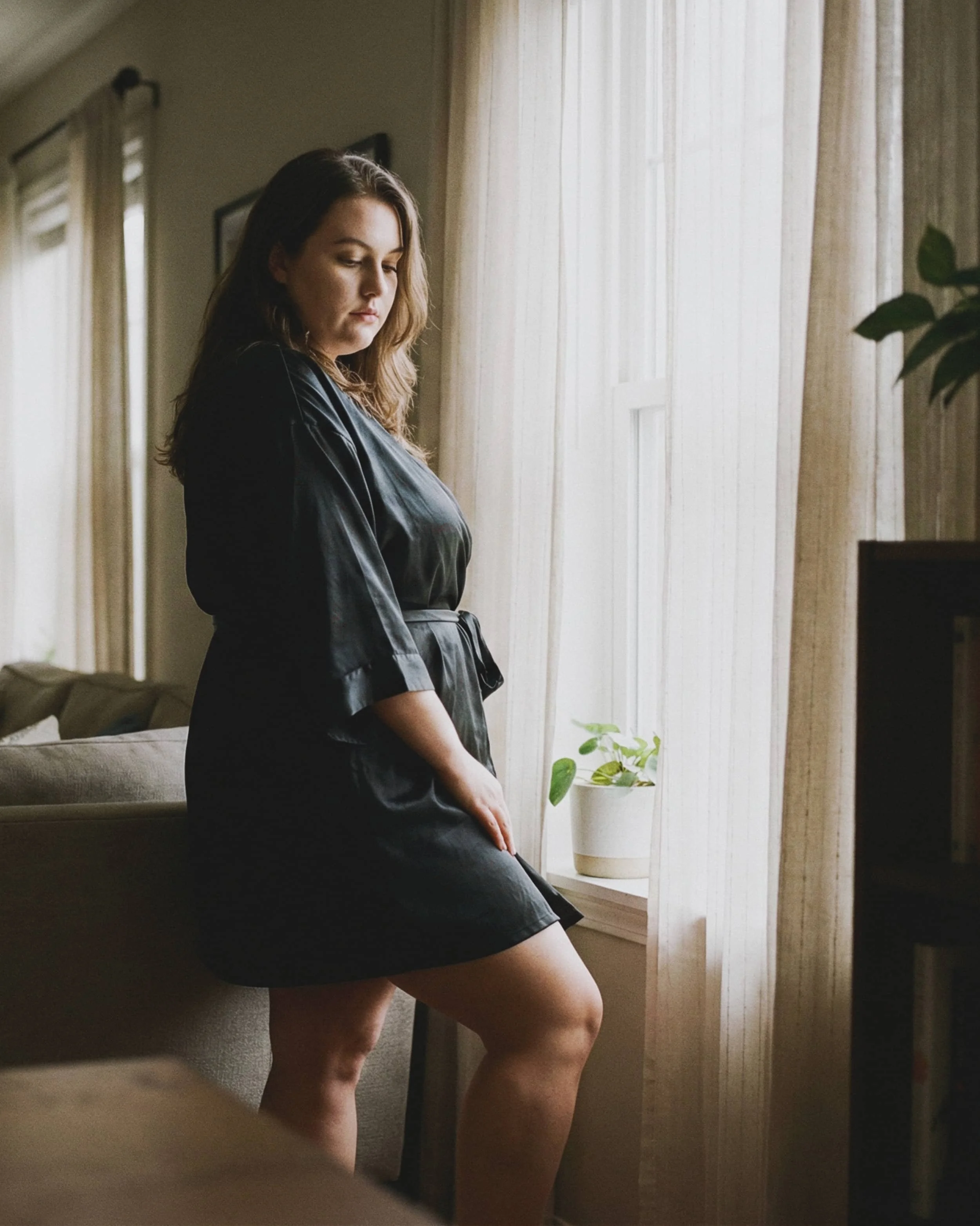 Mujer con bata negra en una habitación, mirando pensativa por una ventana con cortinas claras, junto a una planta en una maceta.