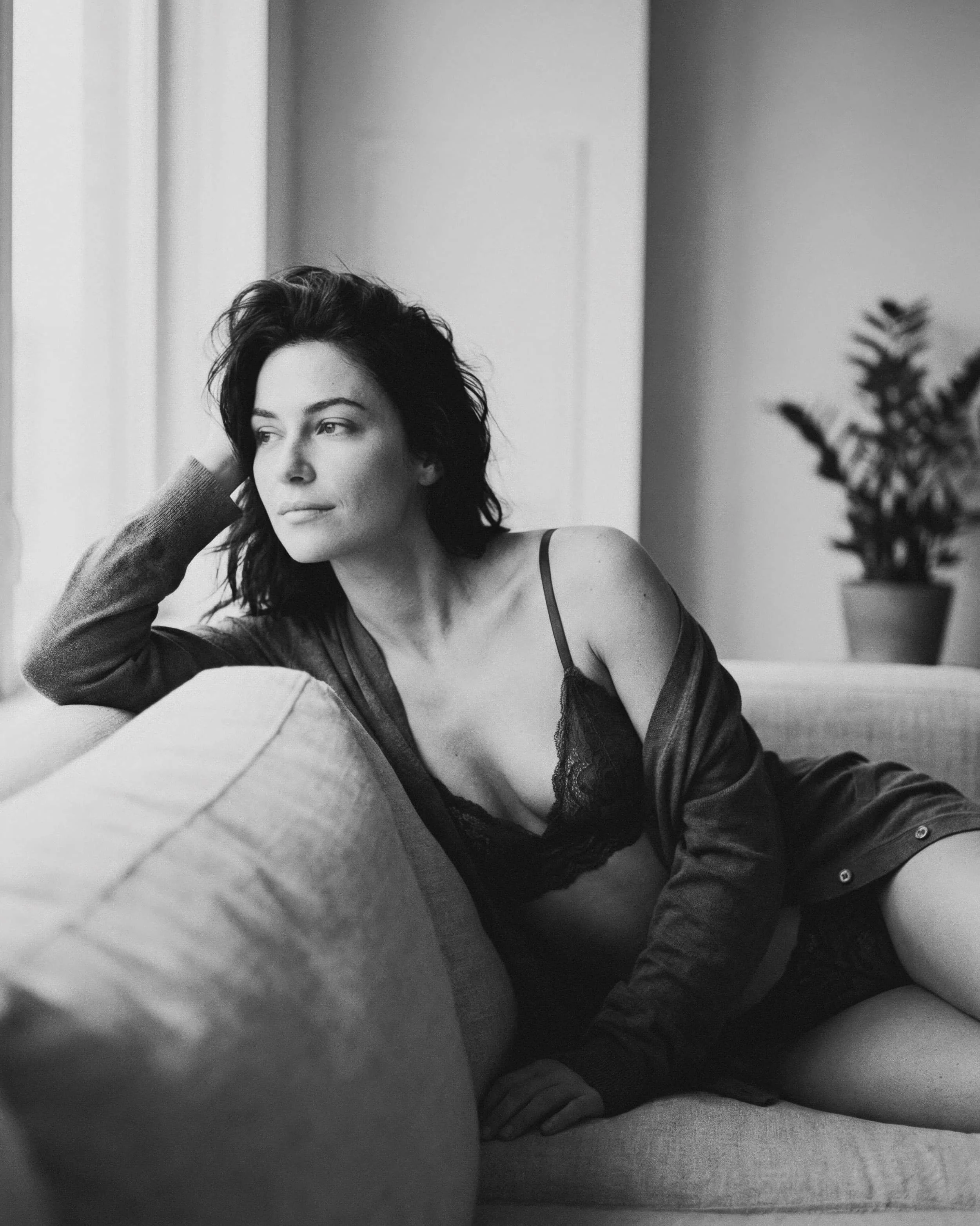 Mujer recostada en un sofá, mirando por la ventana, en ropa interior y un suéter, en un ambiente de interior con planta en el fondo.