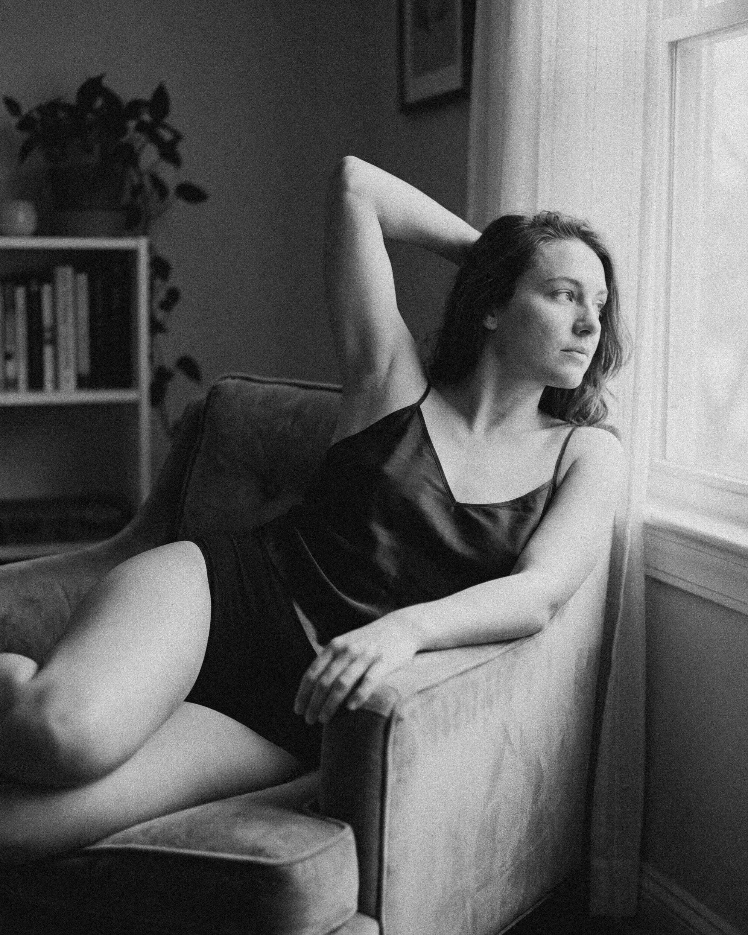 Mujer joven sentada en un sillón, mirando por la ventana, en ropa de seda, en una habitación con luz natural y estantería con libros al fondo.