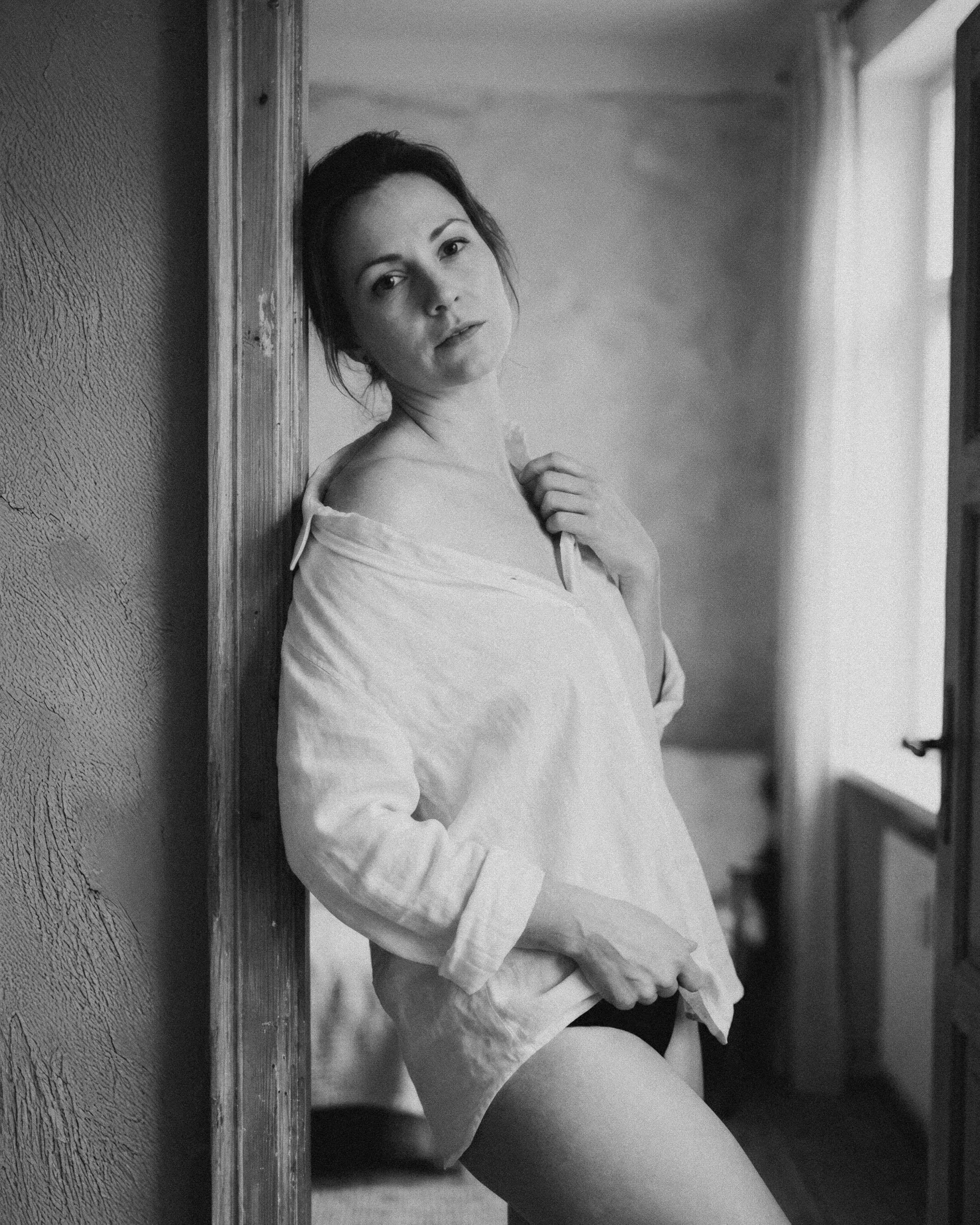 Mujer joven con blusa blanca de manga larga, posando en una habitación, junto a una pared de textura rugosa, con expresión pensativa, en un entorno de interiores con luz natural.