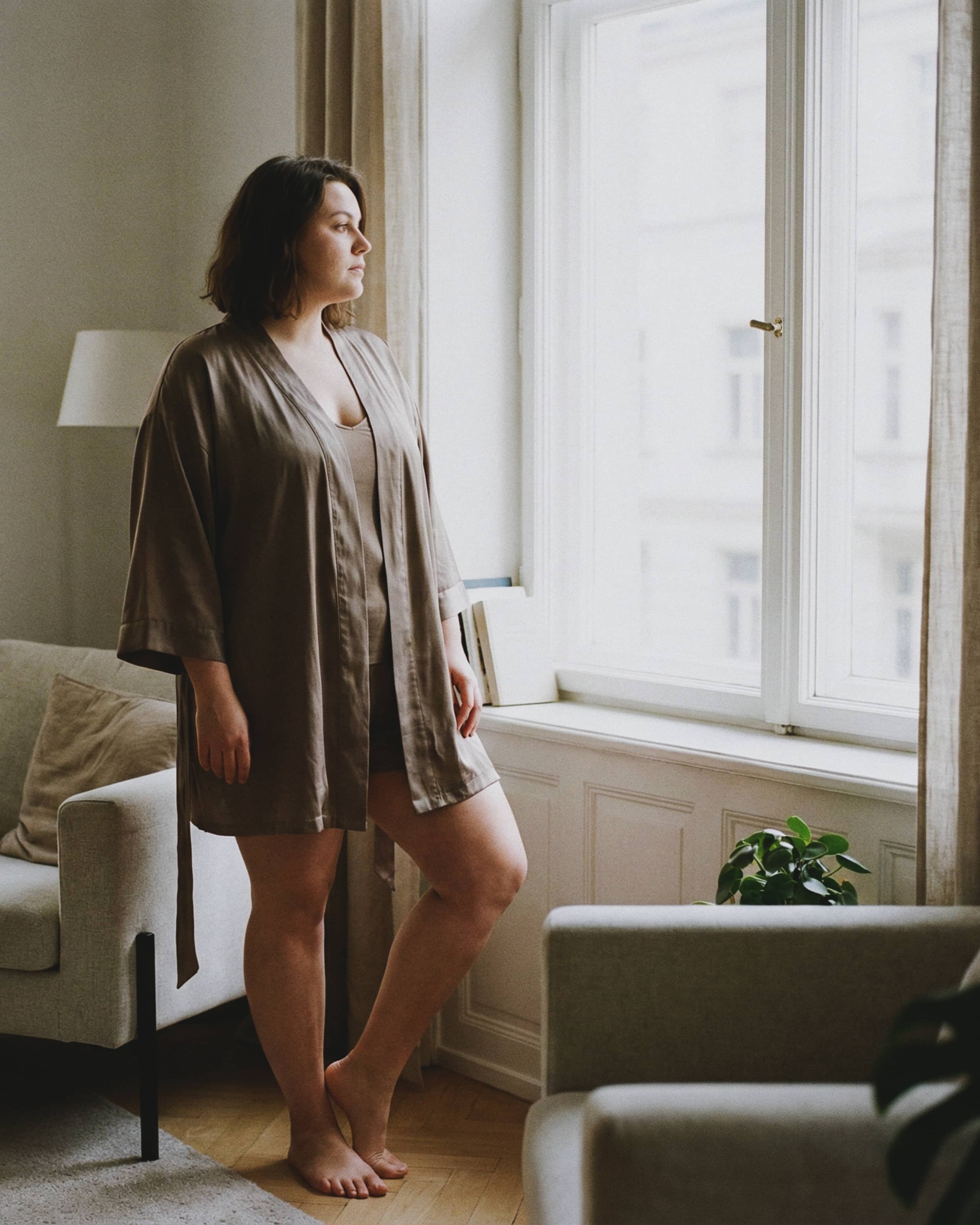 Mujer de cabello castaño oscuro con ropa beige, de pie junto a la ventana observando hacia afuera en un salón con sillones y plantas