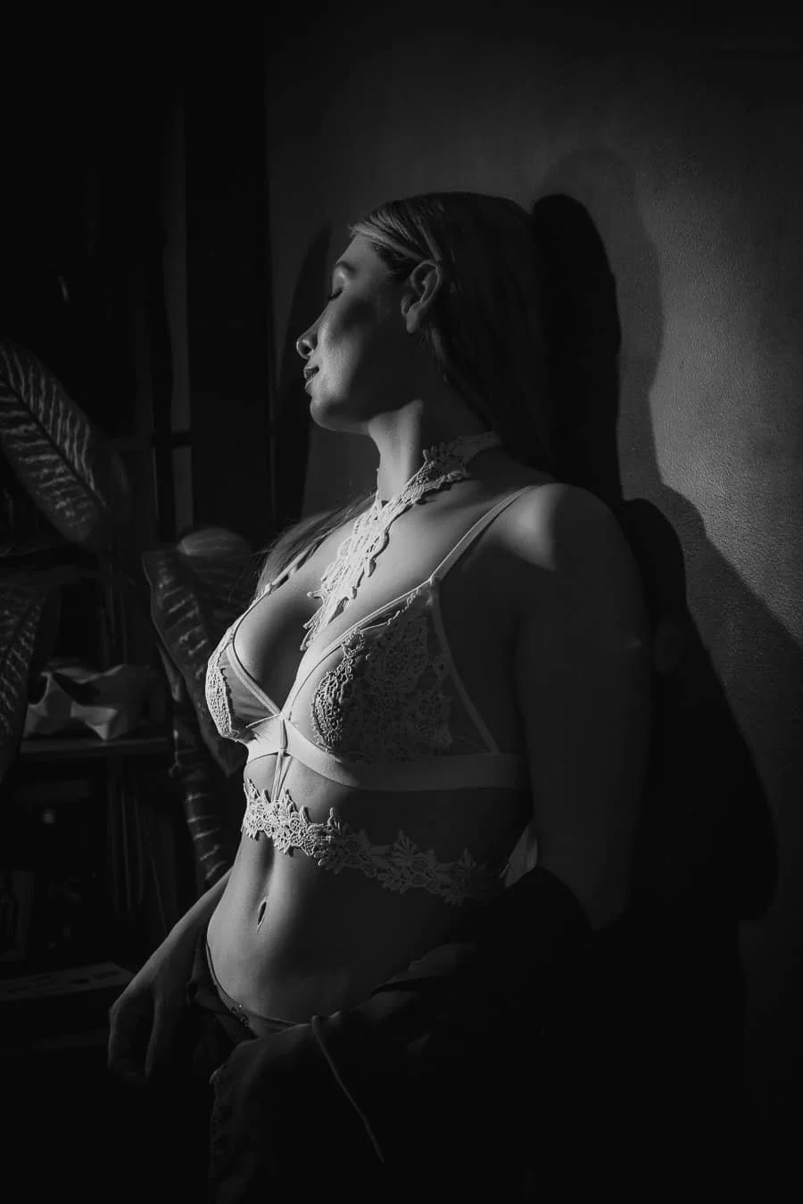 Mujer con cabello liso en ropa de encaje, posando con los ojos cerrados y sombra en un ambiente oscuro.