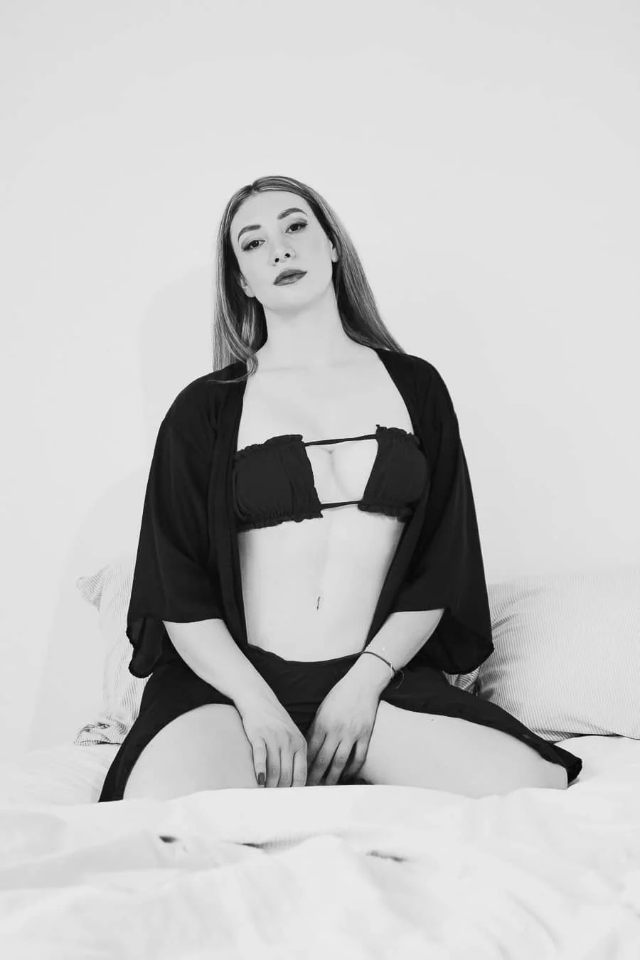 Joven mujer con cabello largo, maquillaje y ropa oscura, sentada en una cama, en una pose frontal, en un entorno minimalista en blanco y negro.