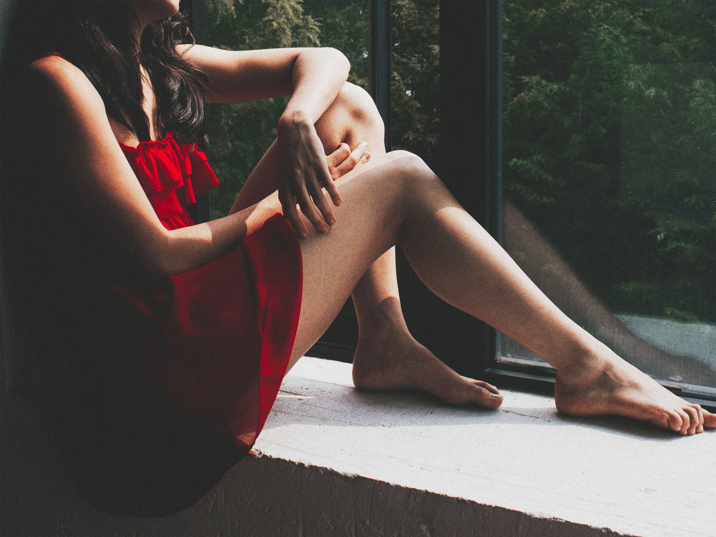 Mujer con vestido rojo sentado en una ventana, con piernas extendidas, mirando hacia afuera, con un fondo de árboles verdes.