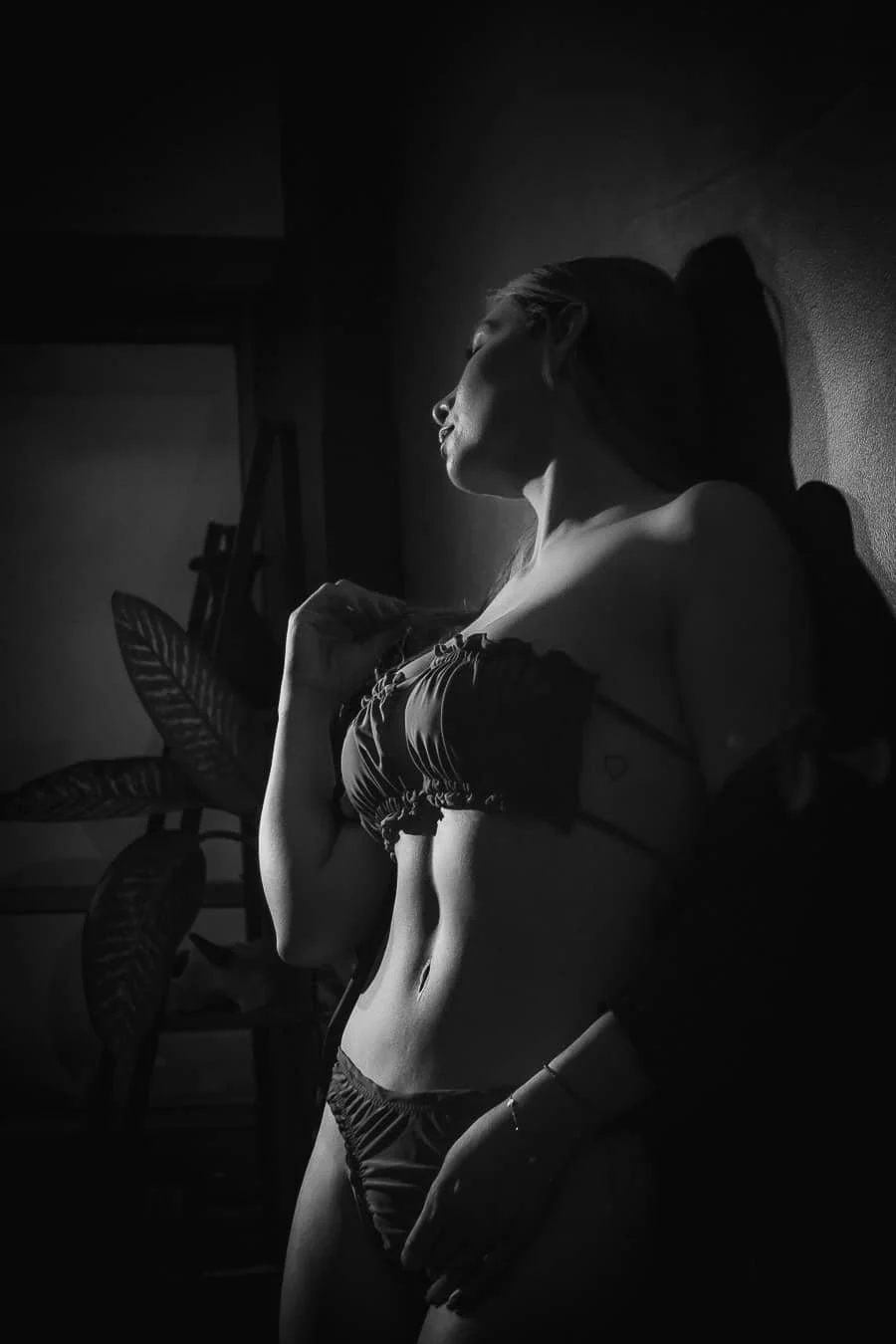 Mujer en ropa interior posando en una pared, iluminada con luz suave en un ambiente oscuro.