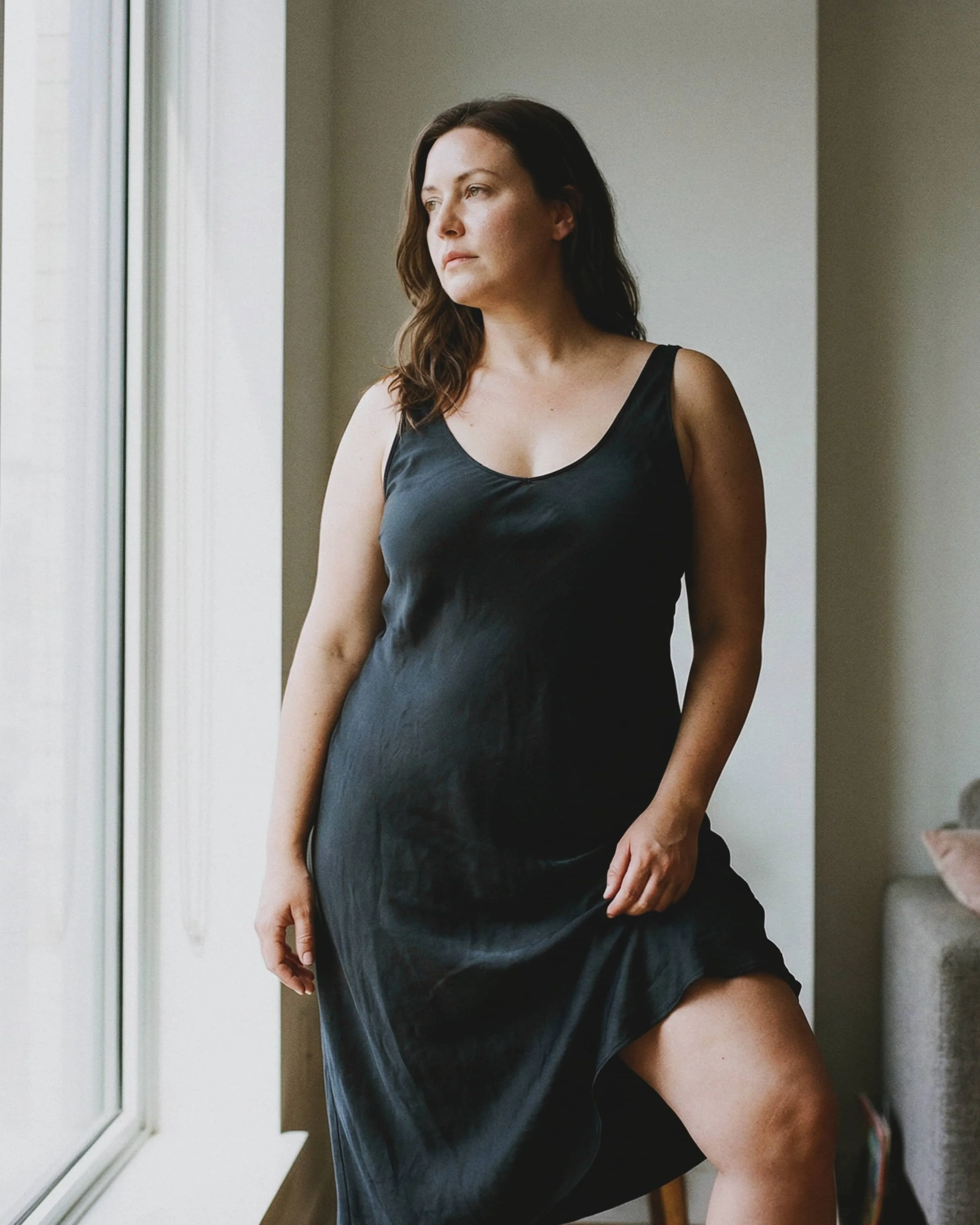 Mujer con cabello castaño oscuro, vestido negro, parada junto a una ventana en interiores