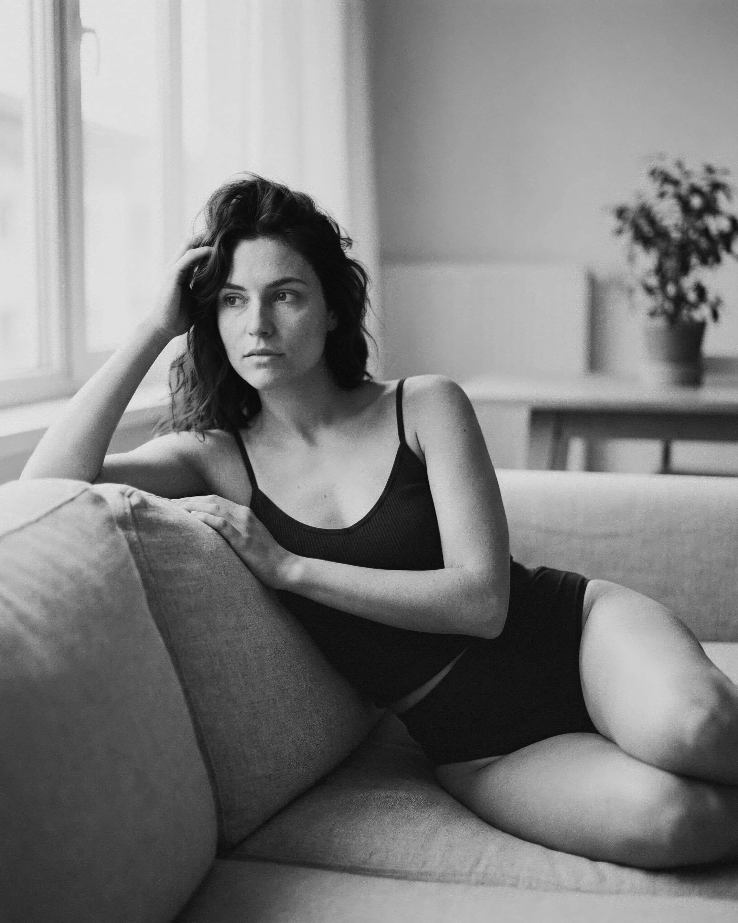 Retrato en blanco y negro de una mujer joven con cabello rizado, sentada en un sofá, con expresión pensativa, en un interior con luz natural y plantas en el fondo.