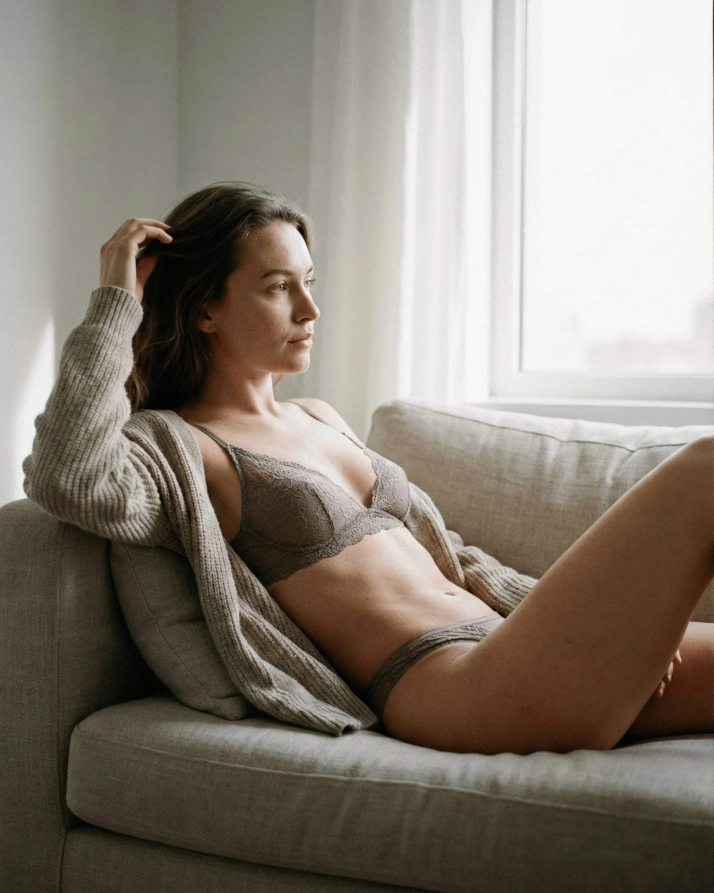Mujer sentada en un sillón, usando ropa interior de encaje y una chaqueta, mirando pensativa por una ventana