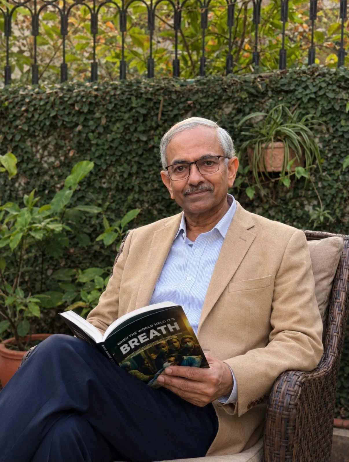 An older man with glasses, wearing a beige blazer and light blue shirt, sitting outdoors in a garden with green plants and a black metal fence, holding a book titled 'When the World Held its Breath'.