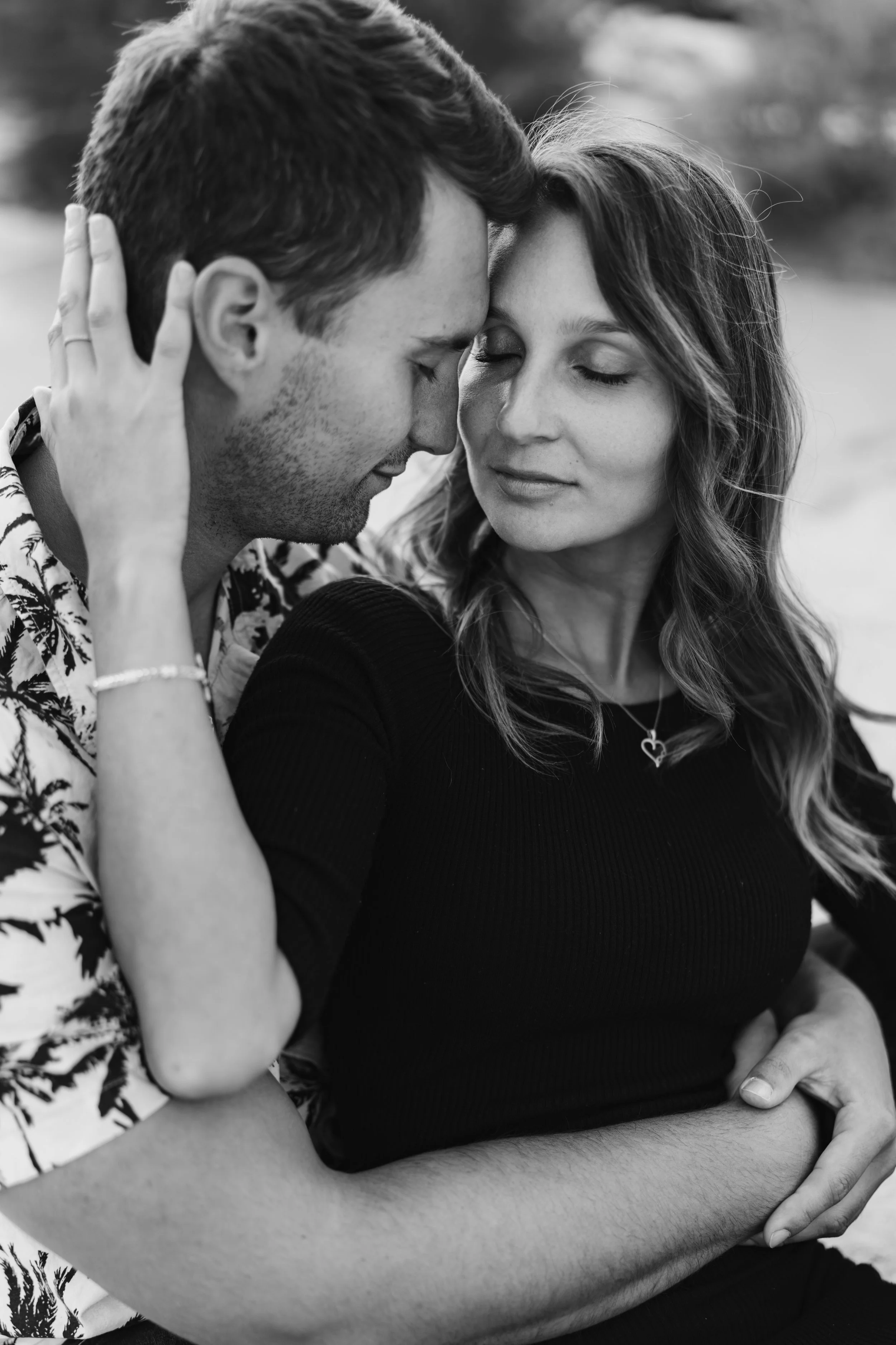 A black and white photo of a couple with their foreheads touching and eyes closed, embracing tenderly outdoors.