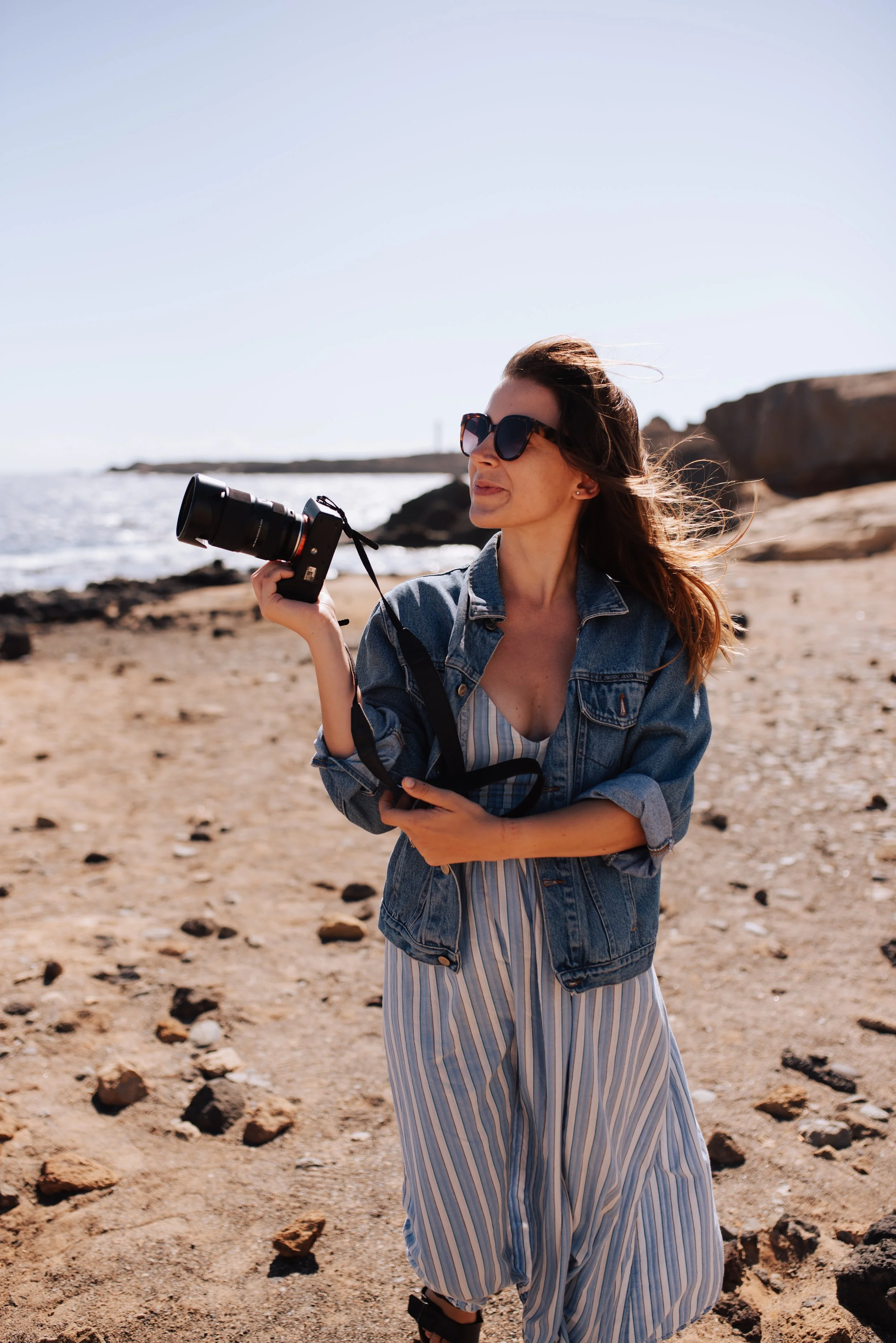 Woman with sunglasses and denim jacket holding a camera on a sandy beach with rocks and the ocean in the background.