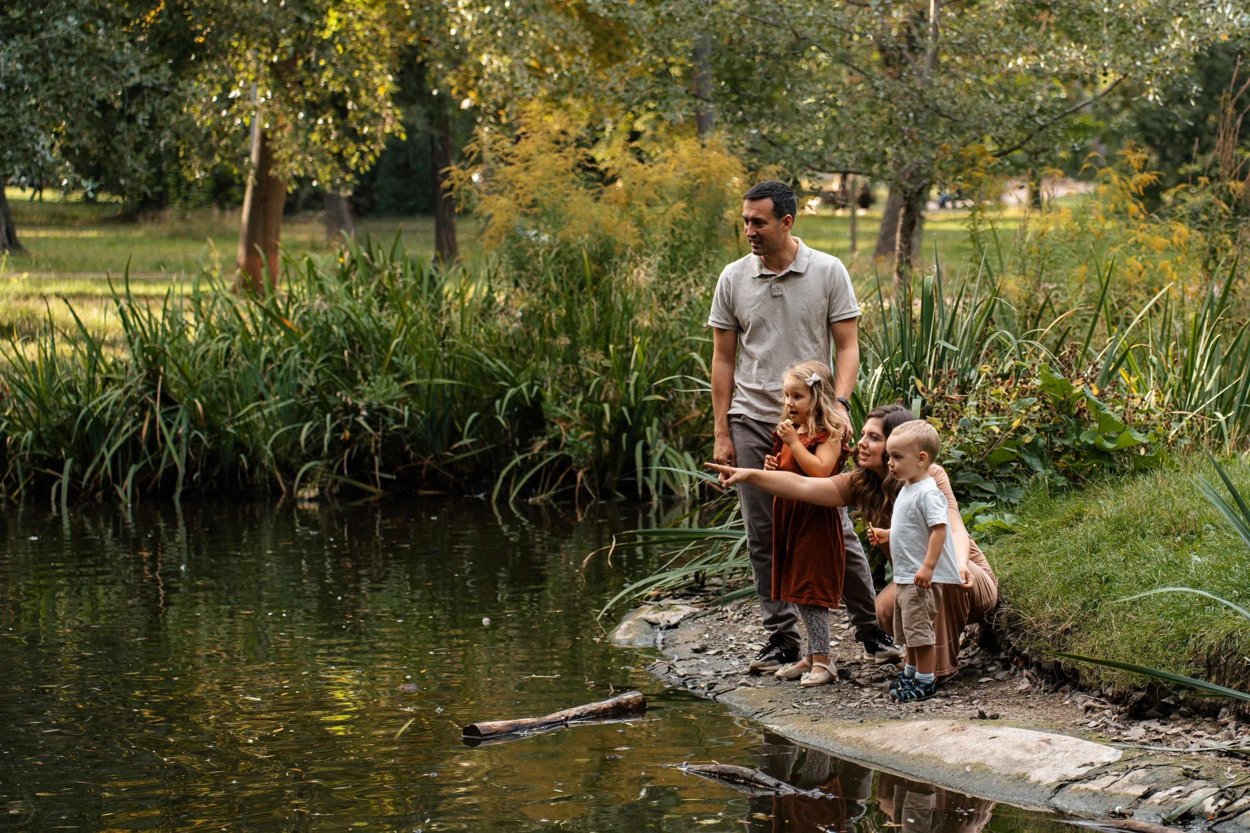 Family of four, including a man, a woman, a young girl, and a young boy, by a pond in a park with trees and tall grass.