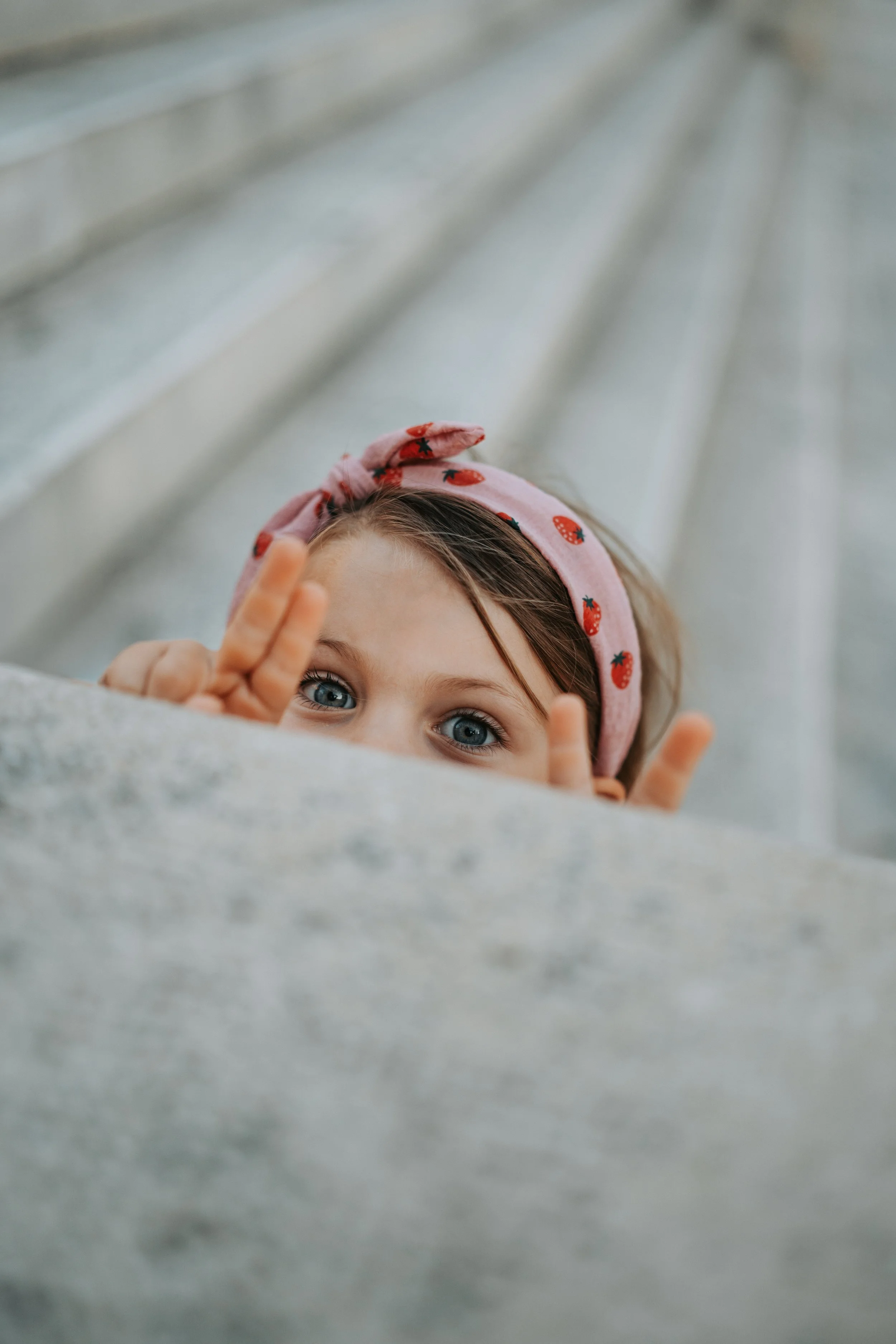 Close-up of a young girl with blue eyes, wearing a pink headband with strawberries, leaning over a gray stone surface, making peace signs with her hands.