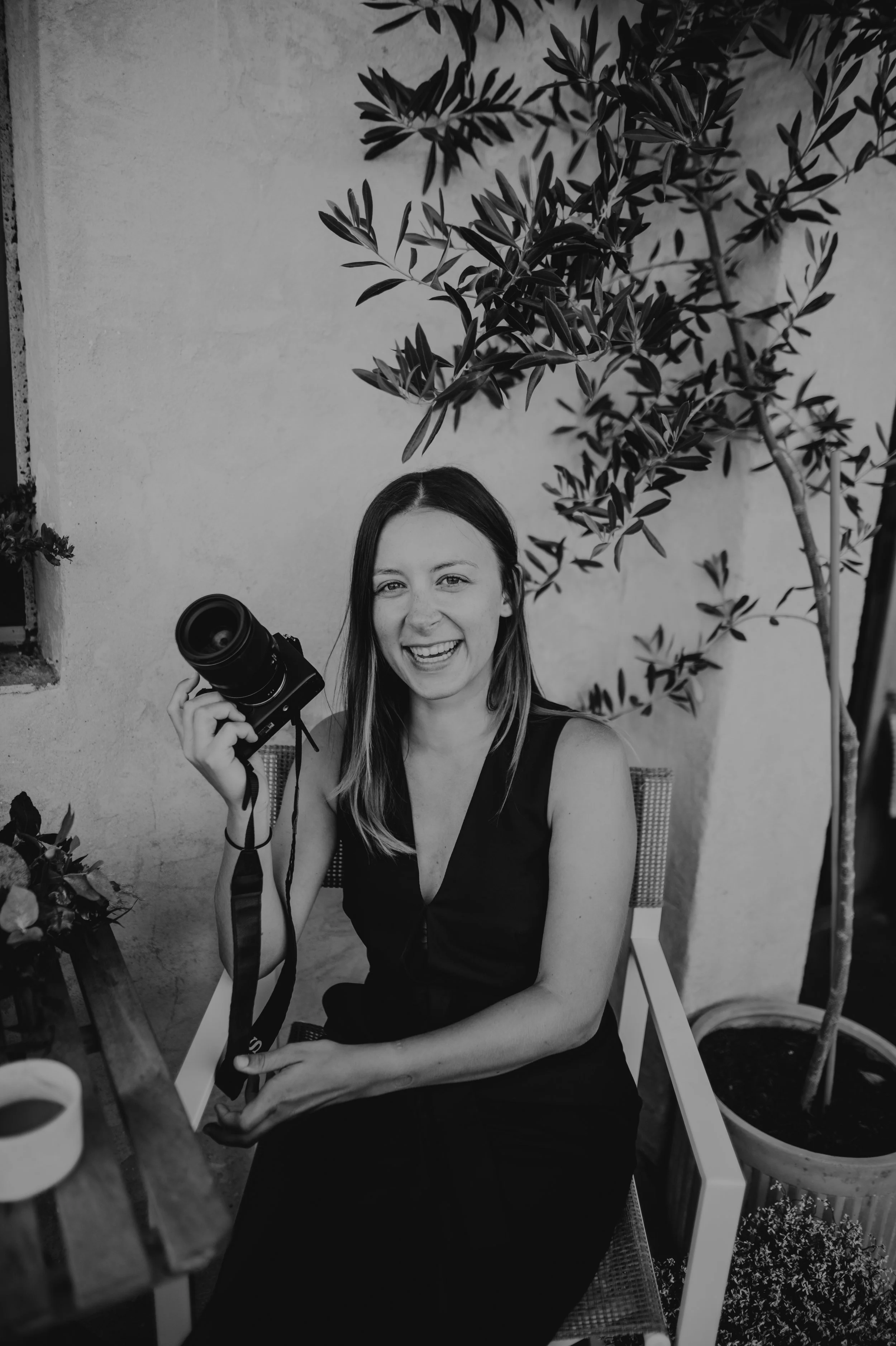 A woman with long hair, wearing a sleeveless black dress, sitting on a chair outdoors, holding a camera in her right hand and smiling at the camera. There are potted plants and a tree behind her.
