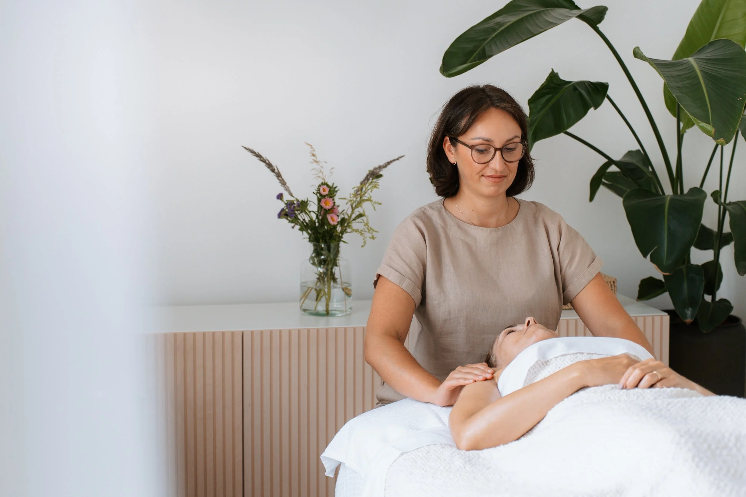 A woman giving a massage to another woman lying on a massage table in a room with large green plants and a vase of flowers.