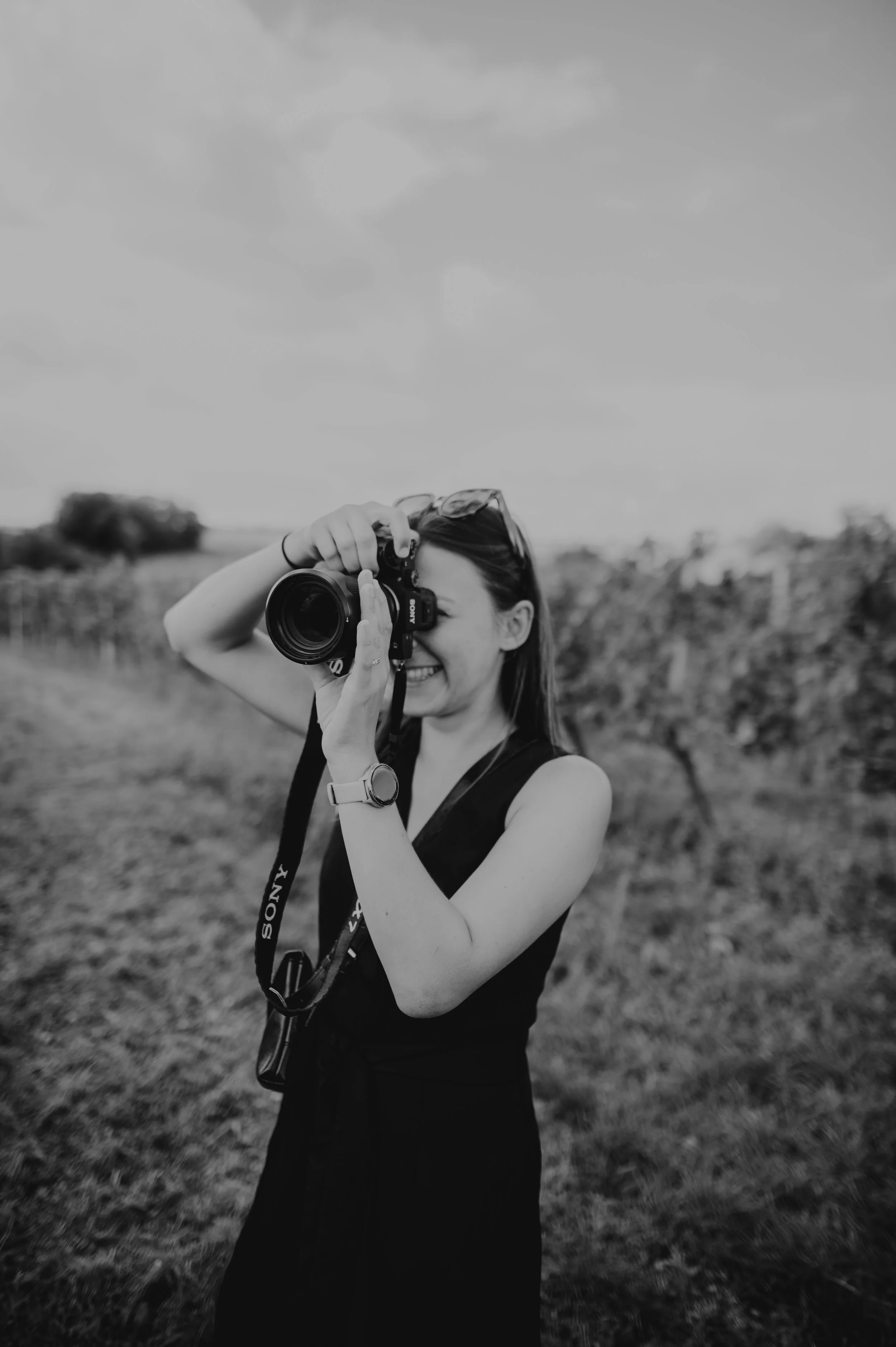 A woman smiling, holding a camera up to her face as she takes a photograph outdoors in a field with rows of plants or vines.