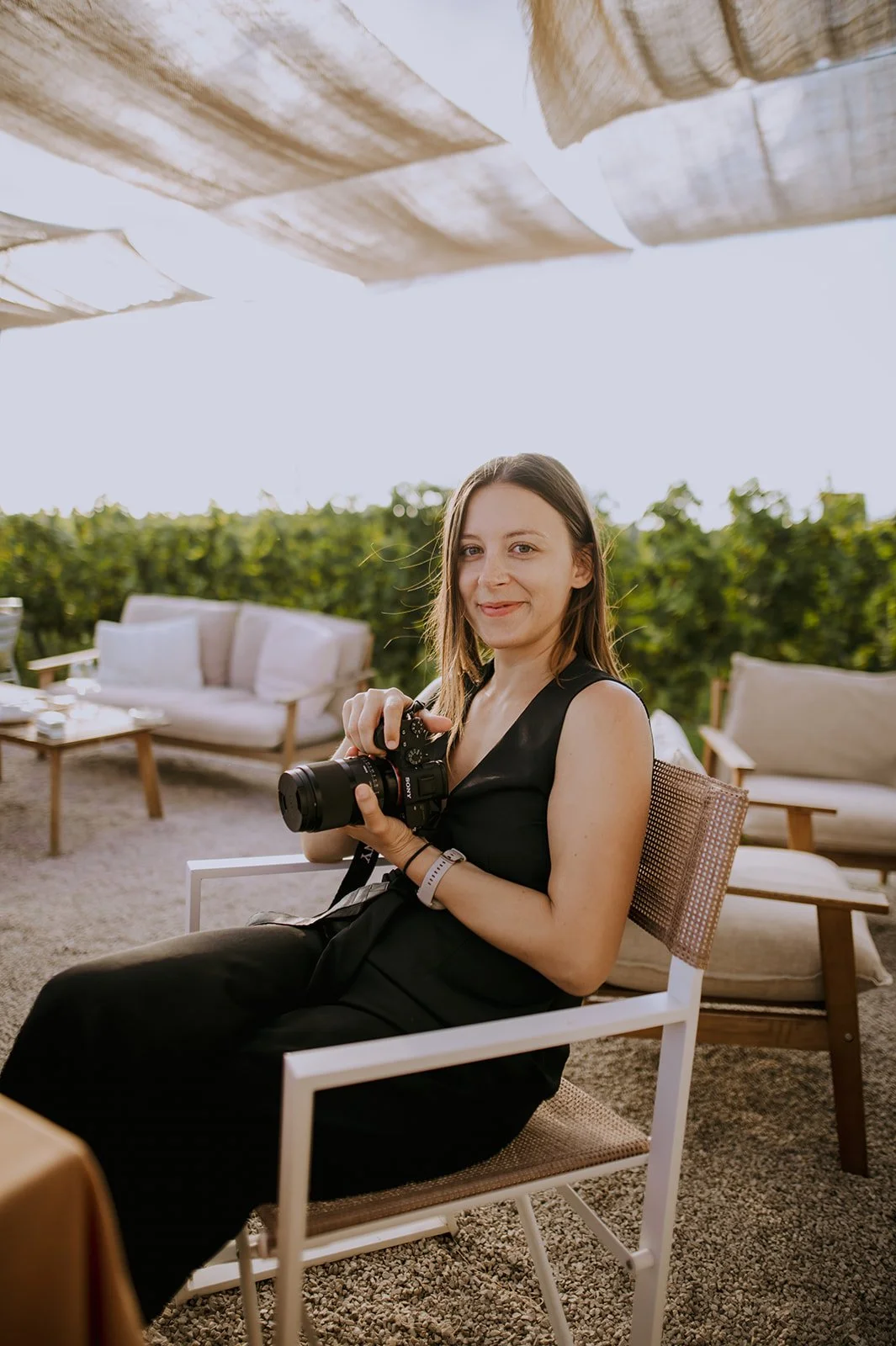 A woman sitting outdoors on a chair, holding a camera, with green shrubbery and outdoor furniture in the background.