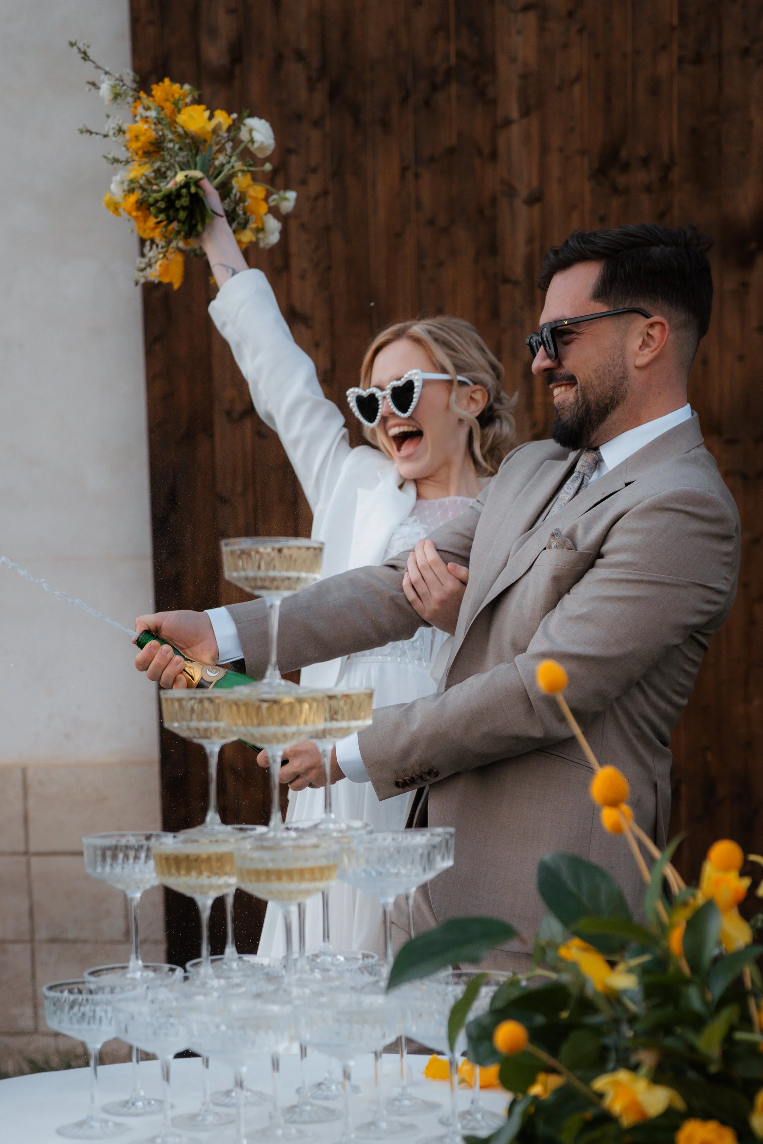 A joyful bride and groom celebrate with a champagne tower at their wedding, with the bride raising a bouquet and both wearing sunglasses.