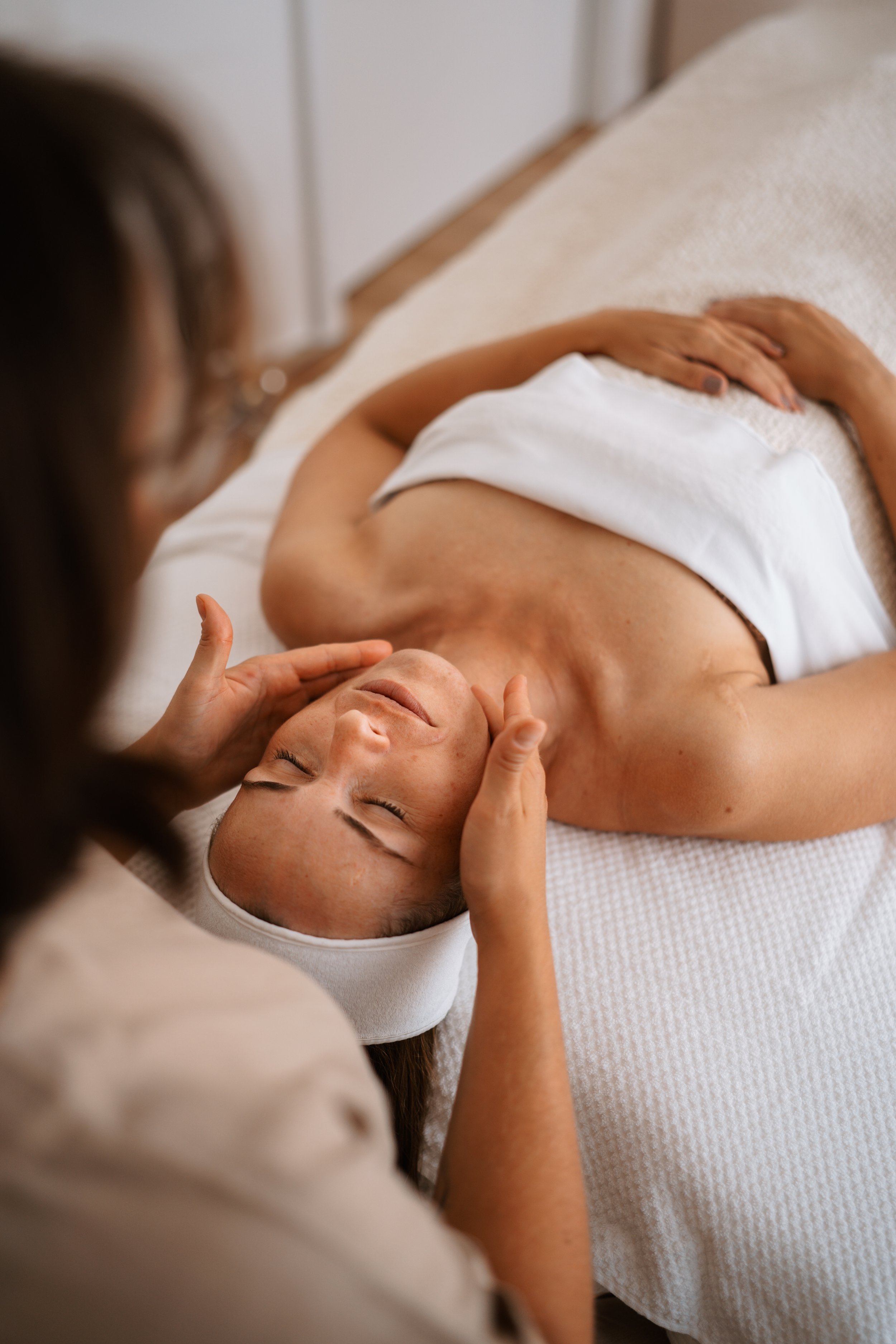 A woman receiving a facial massage from a spa therapist, lying on a spa bed with a towel covering her, in a relaxing spa setting.