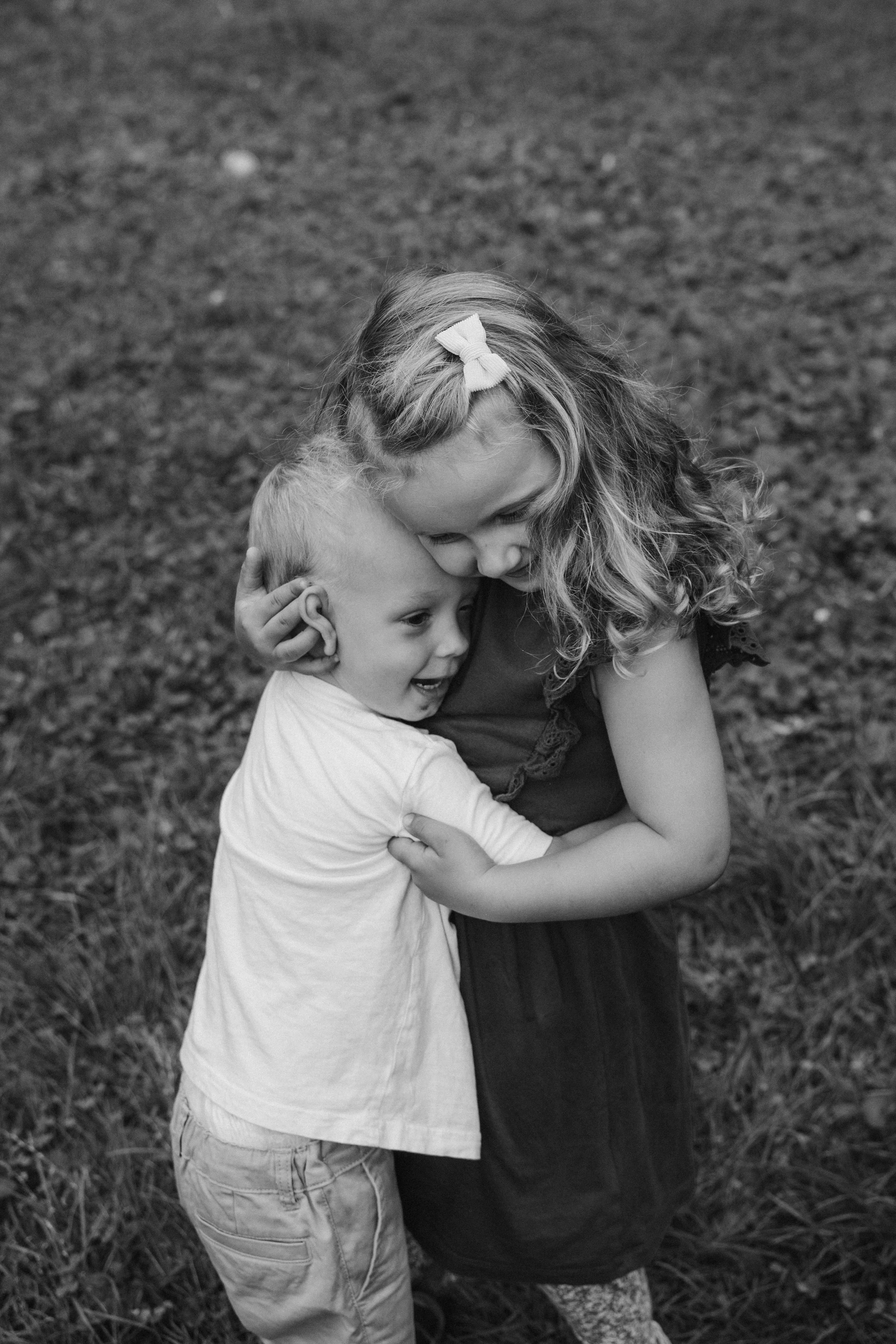 A black and white photograph of a young girl and a boy hugging outdoors on grass.