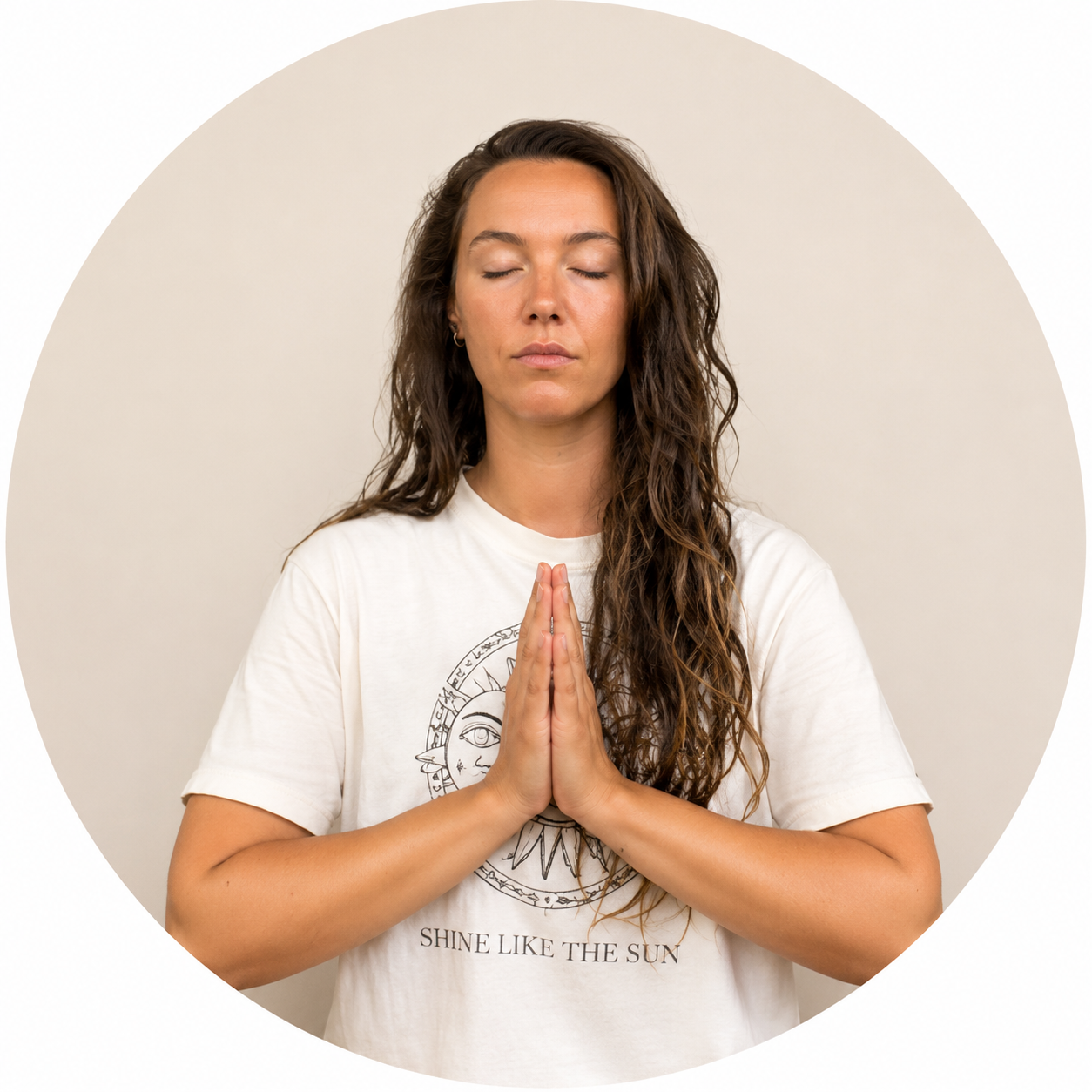 A woman with long wavy brown hair and closed eyes, standing against a plain light-colored background, praying with hands pressed together in front of her chest.