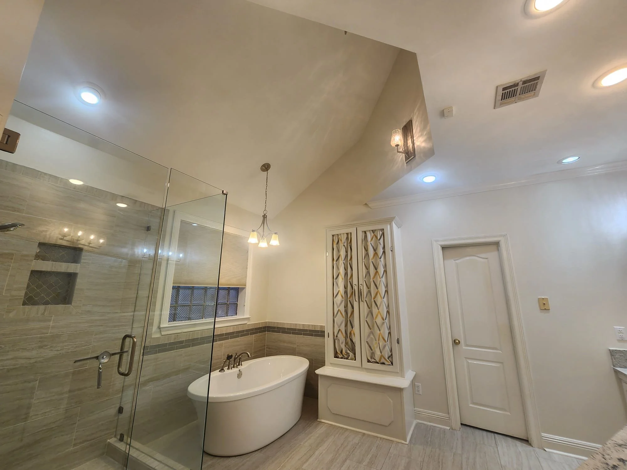 Modern bathroom with a glass-enclosed shower, freestanding bathtub, overhead lighting, and a decorative cabinet with patterned curtains.