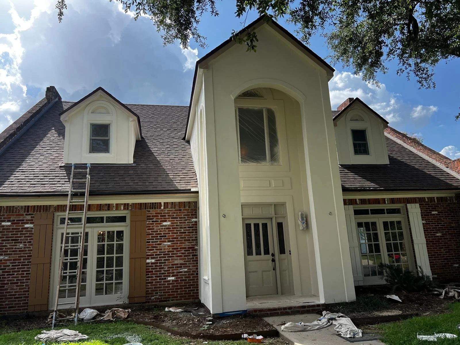 Two-story brick house with white trim, tall center entrance, and dormer windows. A ladder rests against the roof. Paint supplies are on the ground.