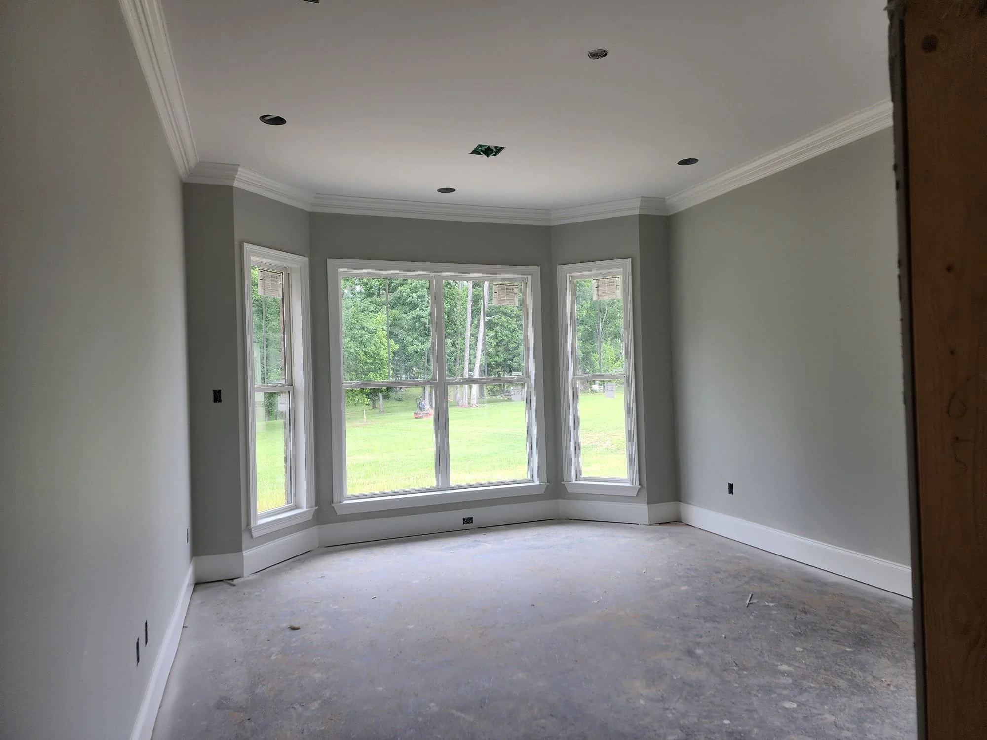 Empty room with gray walls, large bay window overlooking a green lawn, crown molding, and recessed ceiling lights.