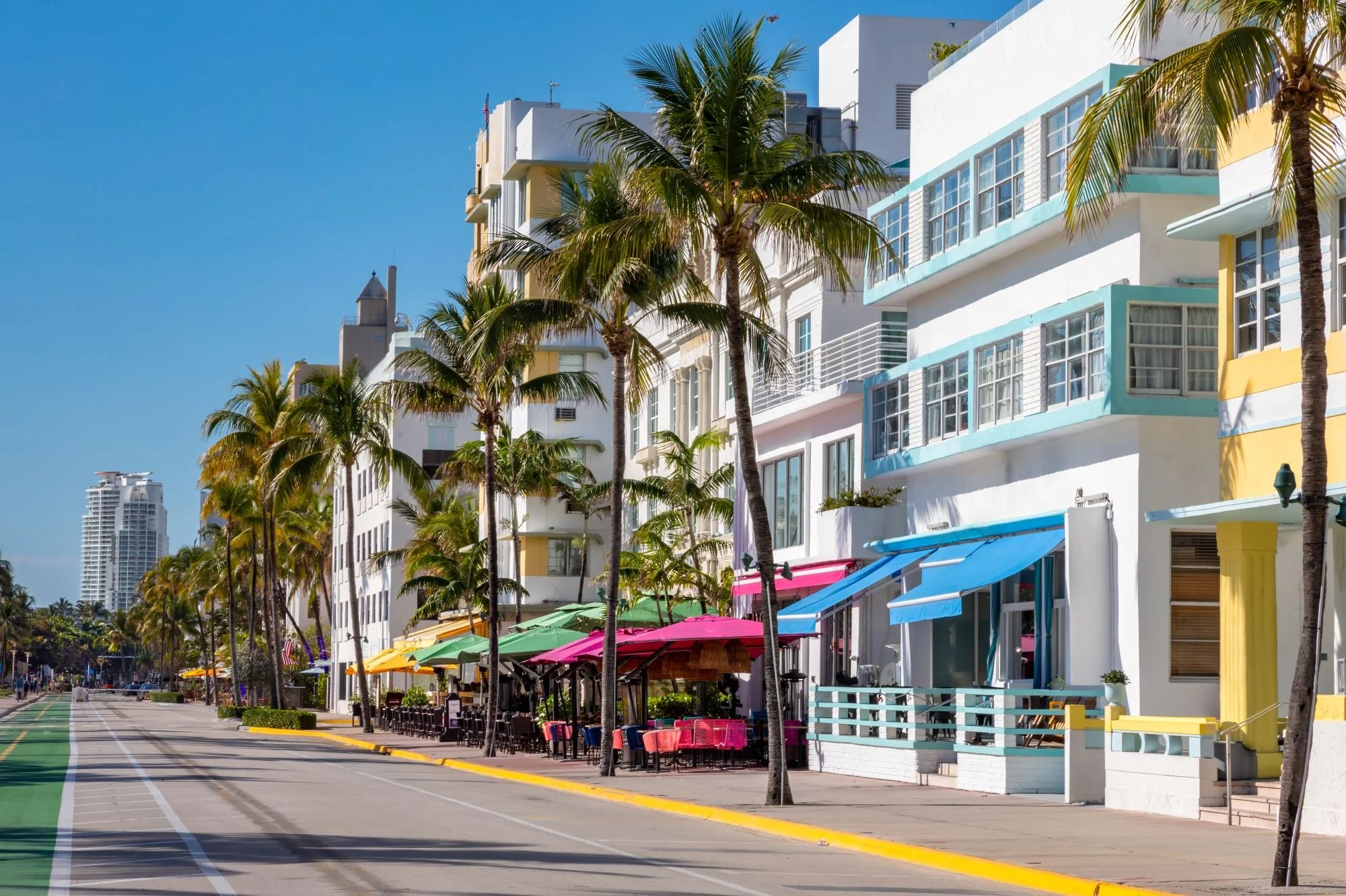 Colorful Art Deco buildings with pastel blue, yellow, and white facades line a street shaded by tall palm trees, with outdoor cafes under pink, green, and blue awnings on a sunny day.