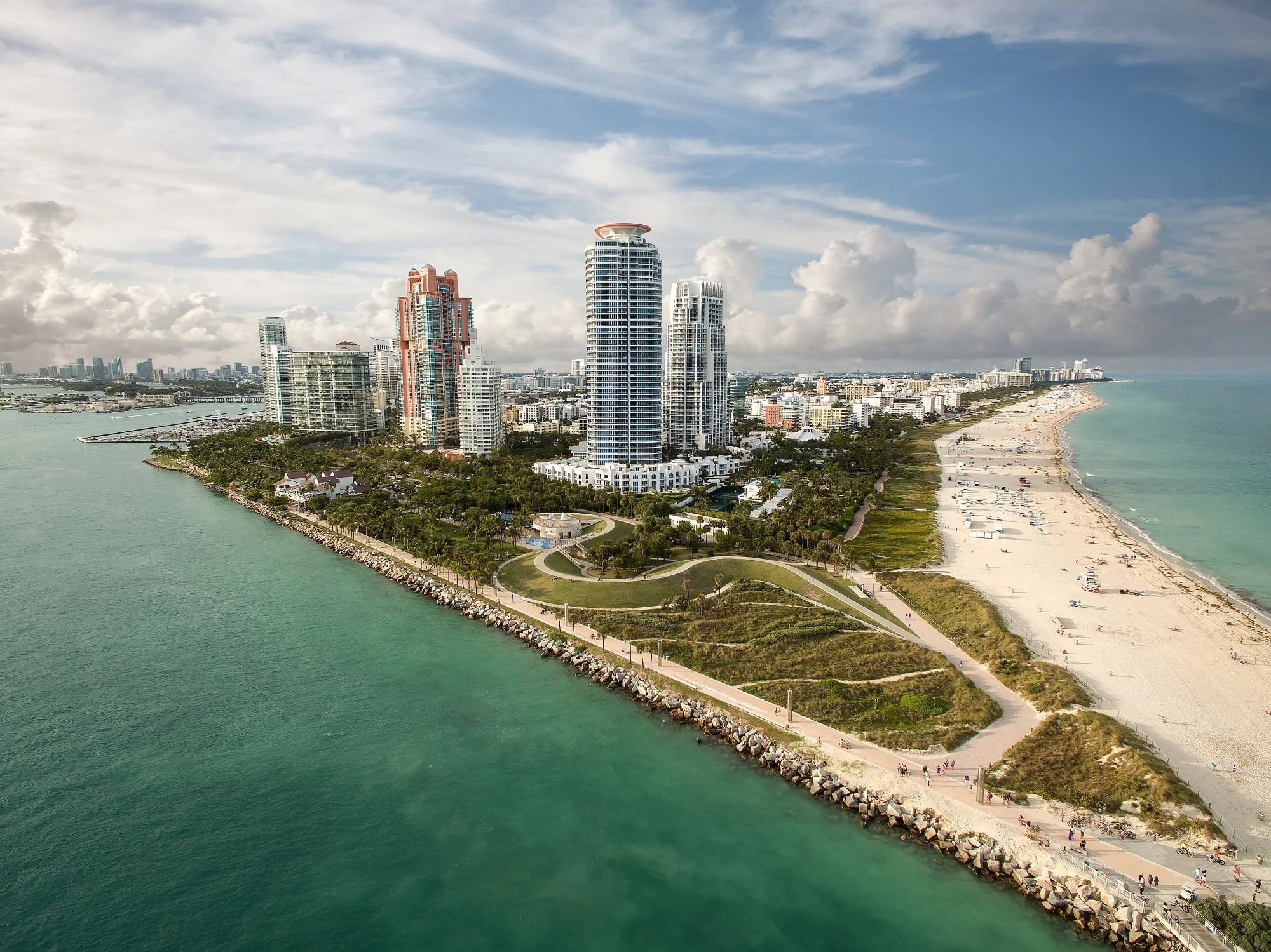 Aerial view of Miami Beach skyline along the coastline, with high-rise buildings, sandy beaches, and ocean, under partly cloudy sky.