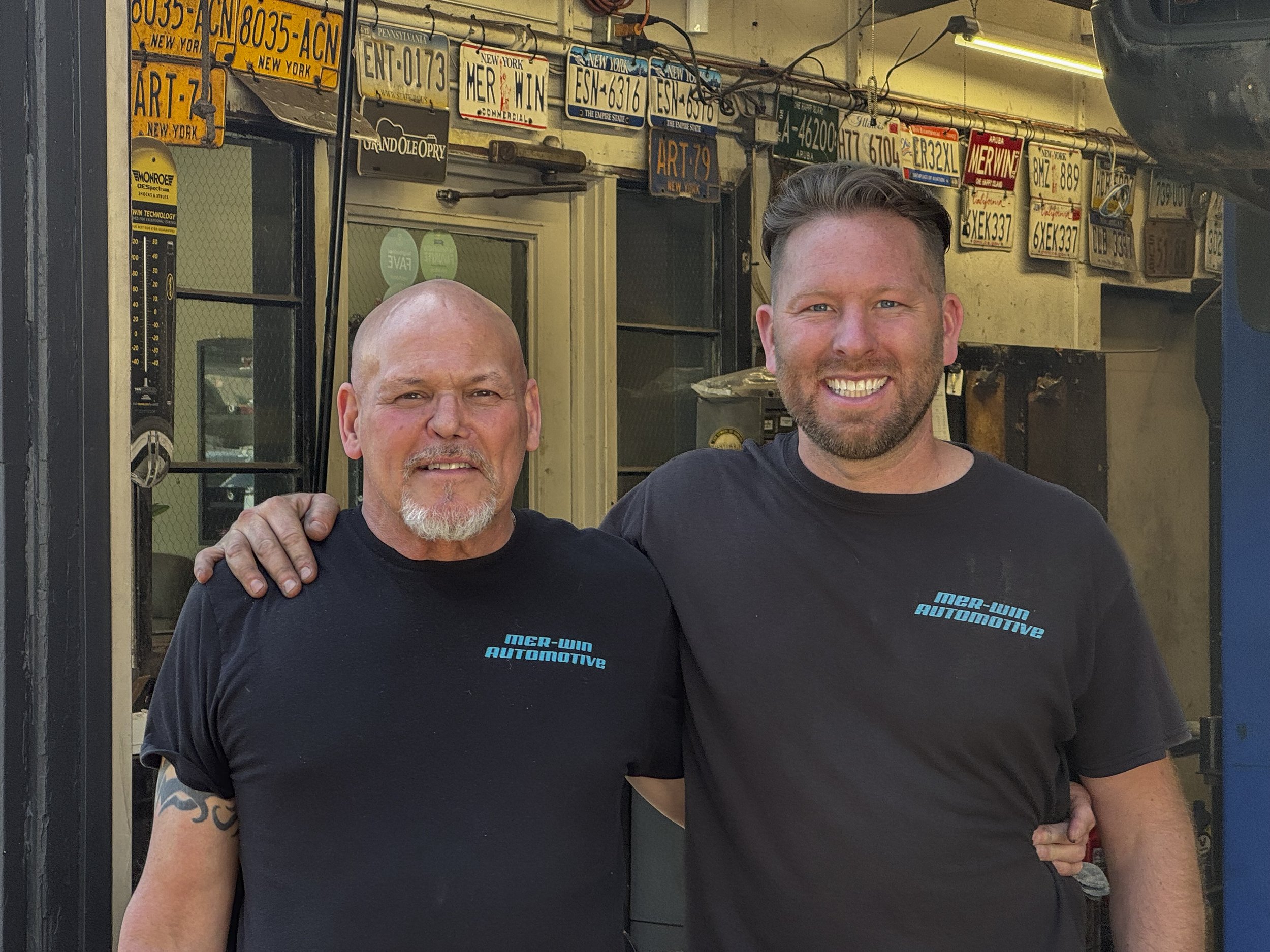 Two men smiling and standing close together outside a building decorated with numerous vintage license plates. One man has a shaved head and a goatee, and the other has short brown hair and a beard.