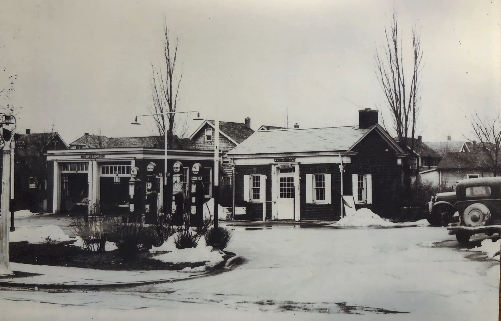 A black and white photograph of a vintage gas station with multiple fuel pumps, a small brick building with large windows, and parked cars from the early to mid-20th century. Snow is on the ground, and leafless trees are in the background.