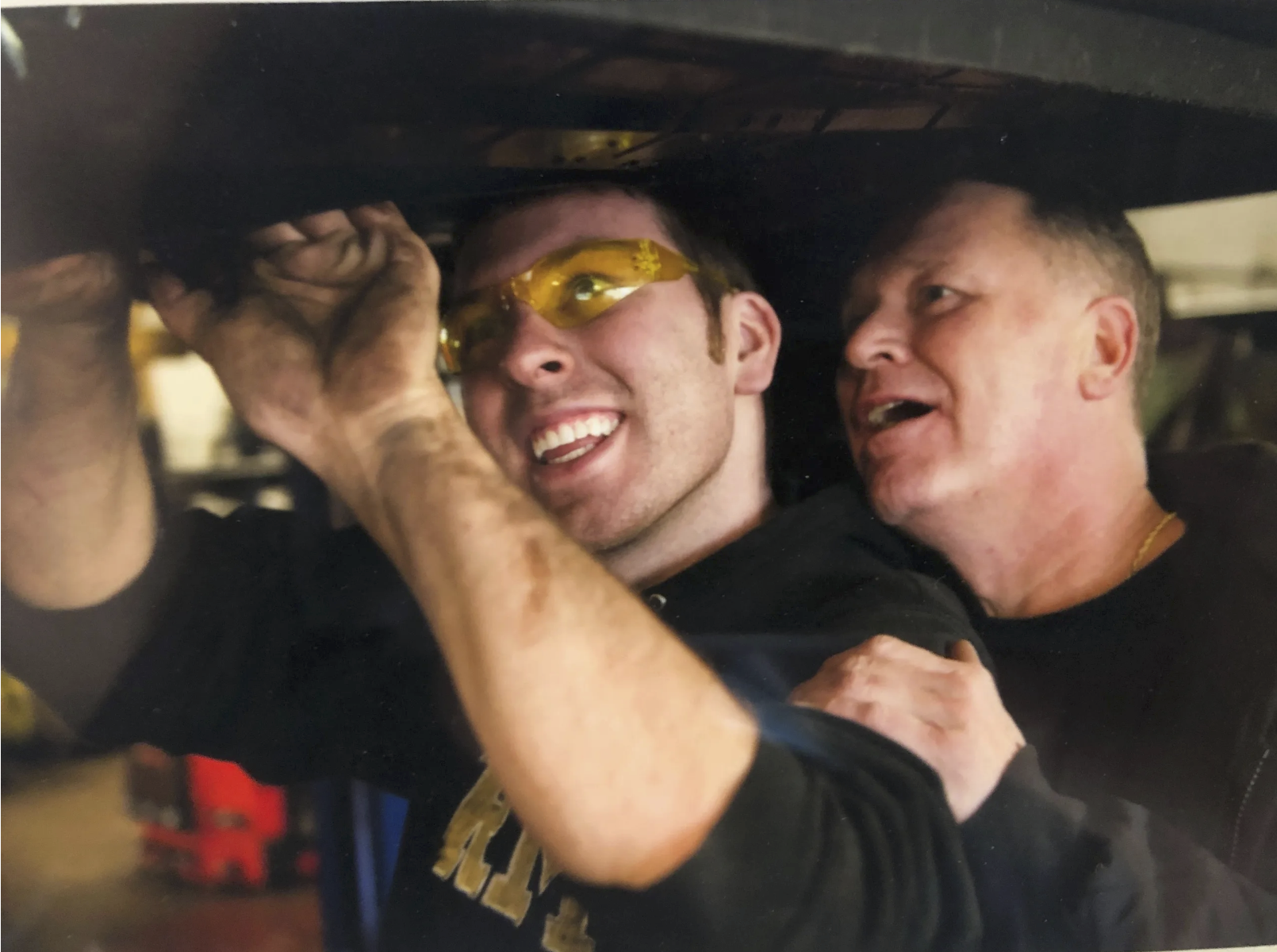 Two men with short hair, one wearing yellow safety glasses, appear to be working together under a vehicle, smiling and engaged in a mechanic task.